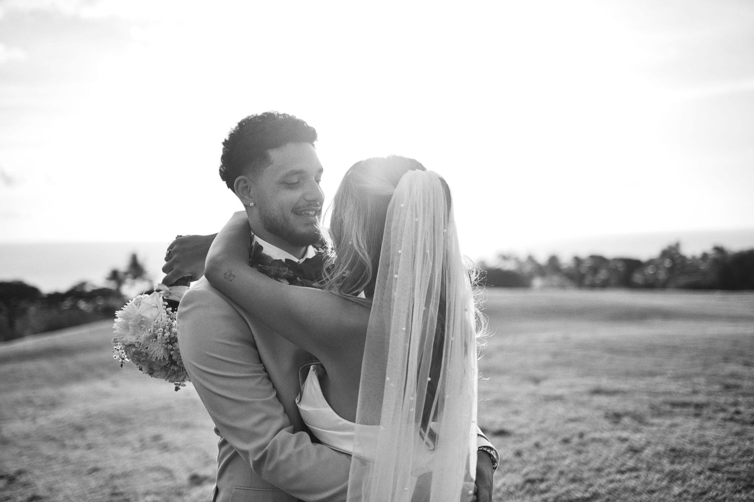 Black and white photo of a groom lifting his bride in a field, her veil flowing, with a scenic outdoor backdrop.