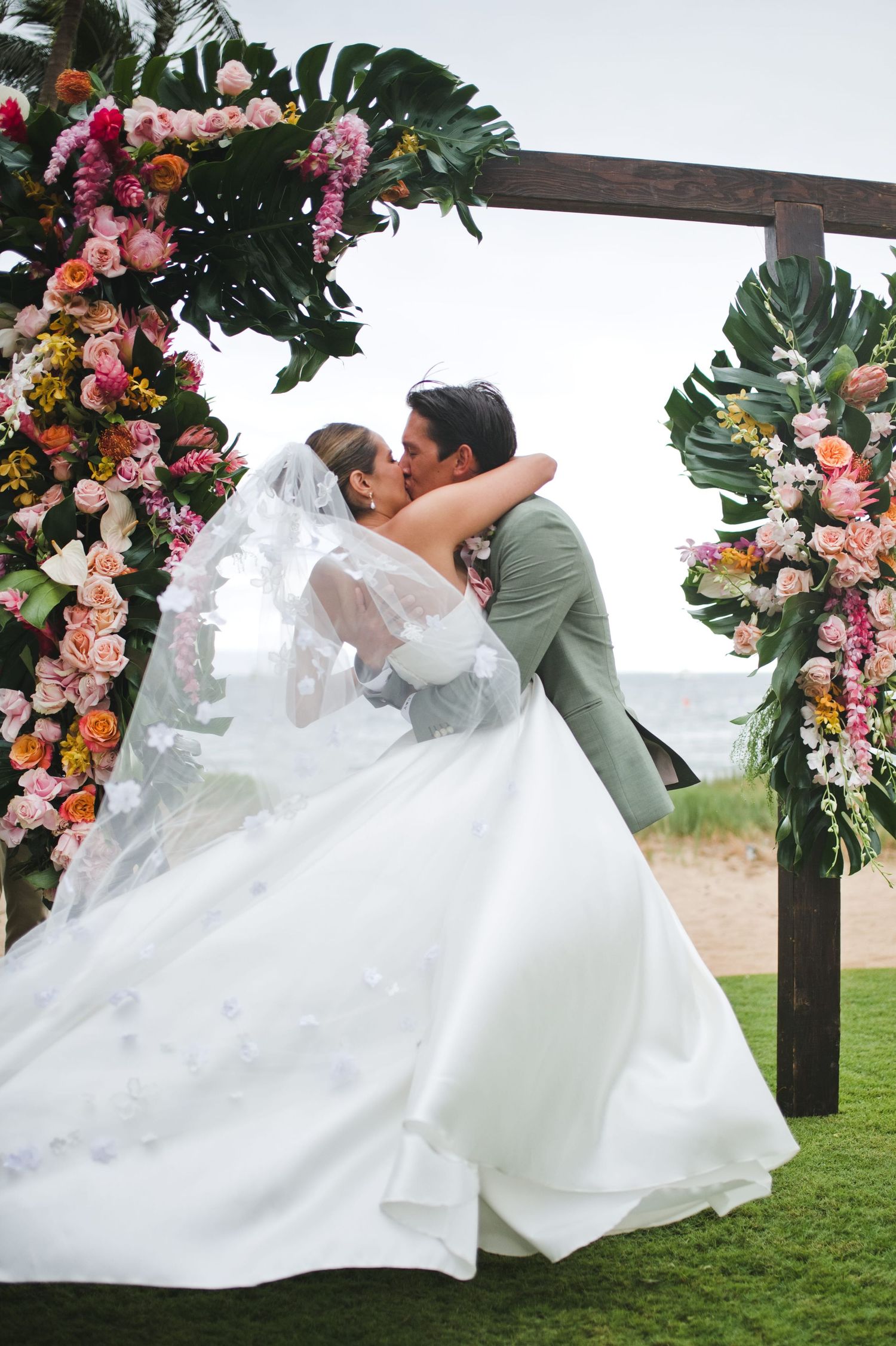Bride and groom share a romantic kiss under a tropical floral arch with monstera leaves on a beach wedding ceremony.