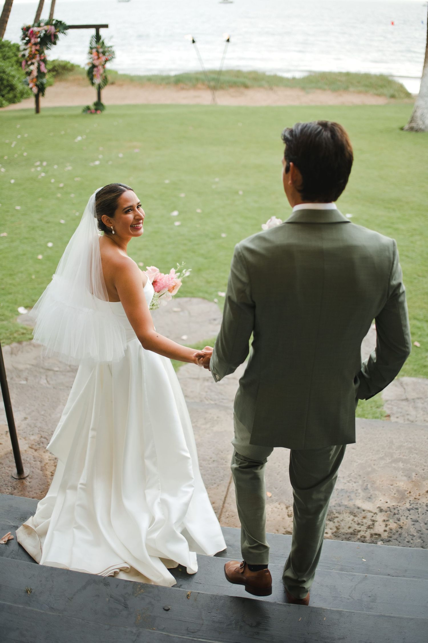 Bride in white gown and veil holds hands with groom in olive suit at outdoor beach wedding ceremony.