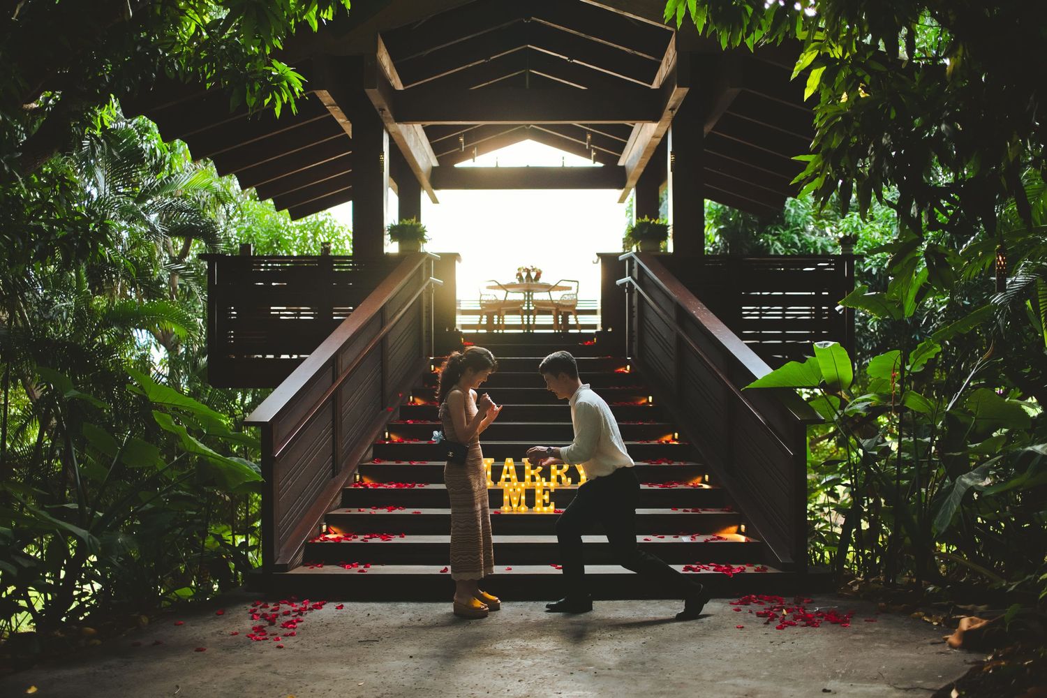 Romantic marriage proposal on tropical bridge stairs with glowing MARRY ME sign surrounded by lush greenery at sunset.
