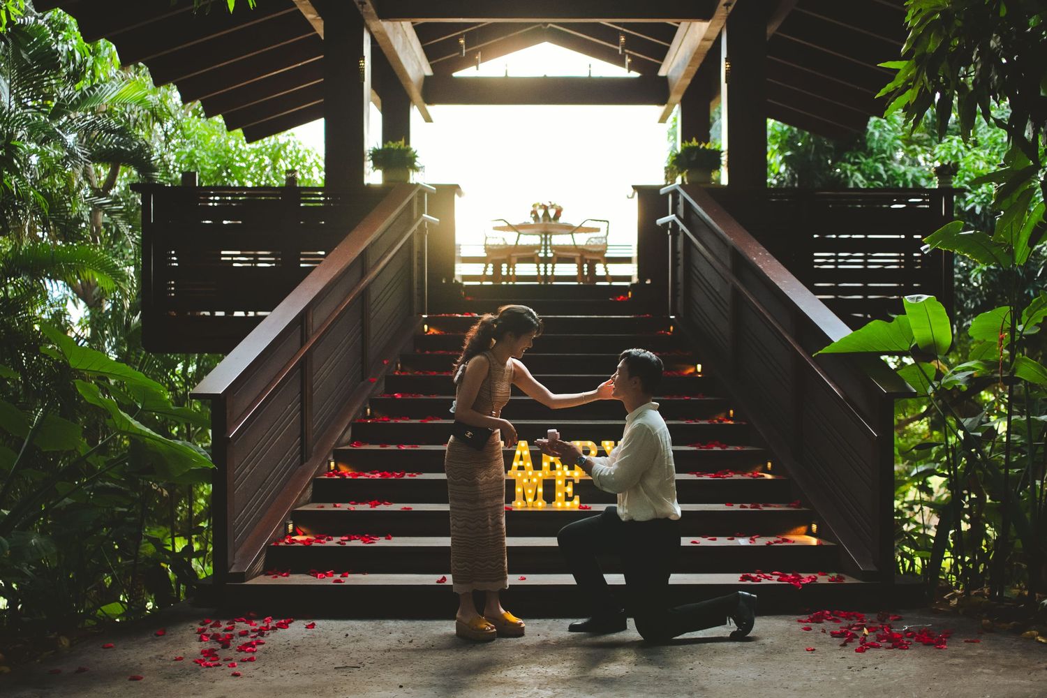 Man proposes to woman on flower-petal-lined stairs at a tropical resort gazebo at sunset.