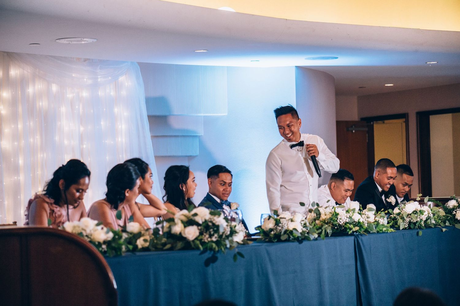 Wedding party seated at an elegant head table decorated with white floral arrangements and soft lighting.