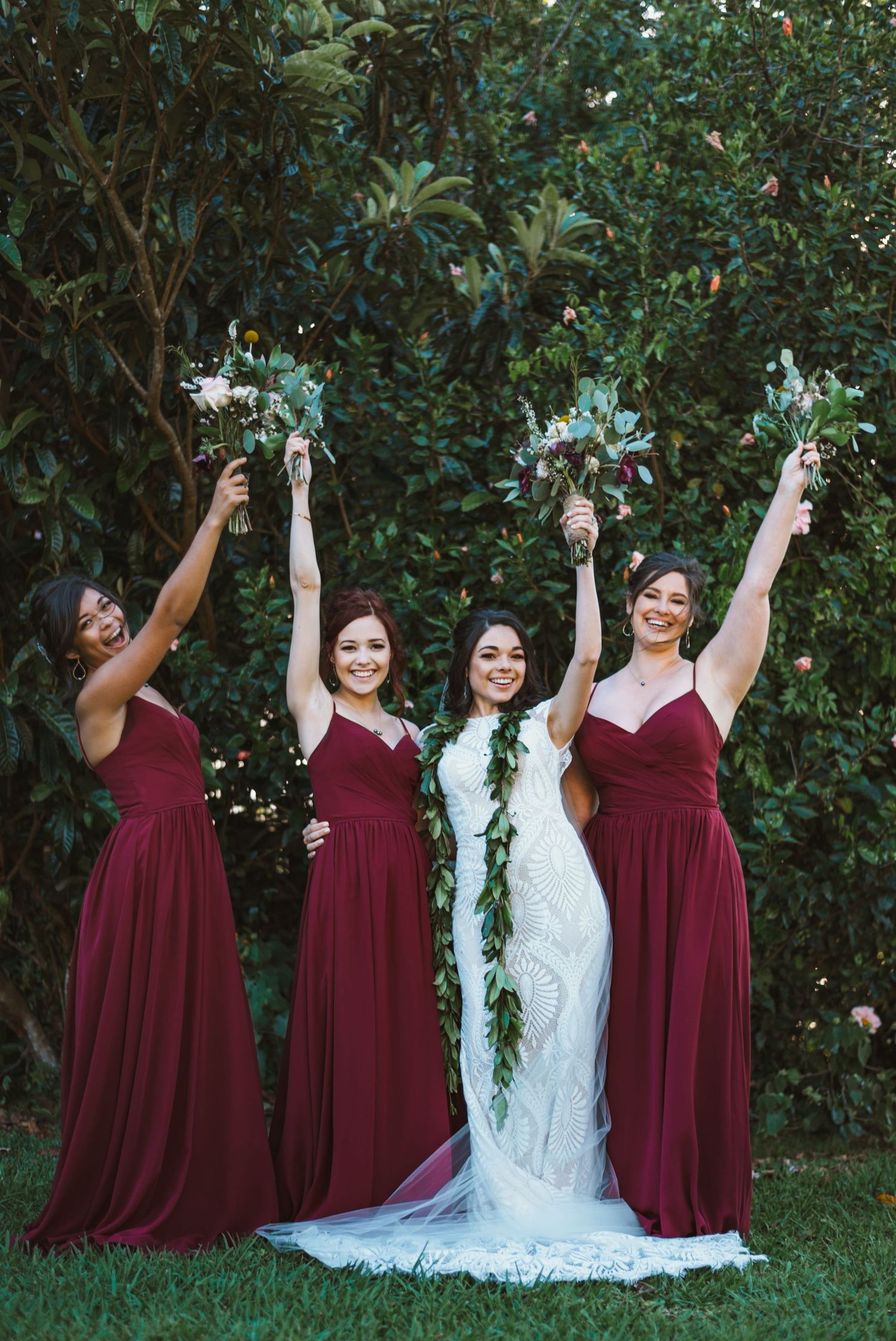 Bride in white lace gown with bridesmaids in burgundy dresses raising bouquets outdoors.