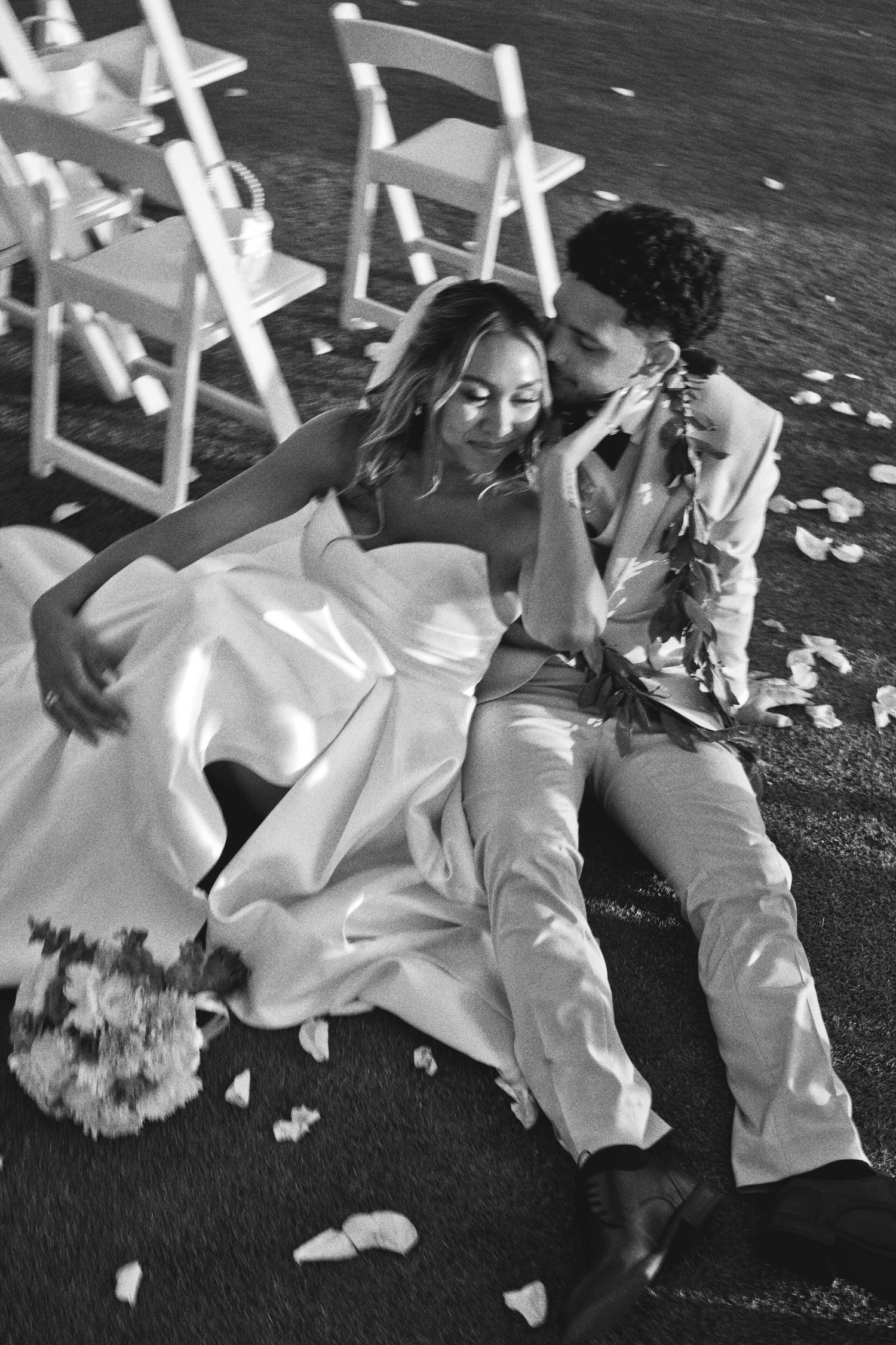 Black and white photo of a bride and groom sitting on the ground surrounded by flower petals and white chairs at their wedding.
