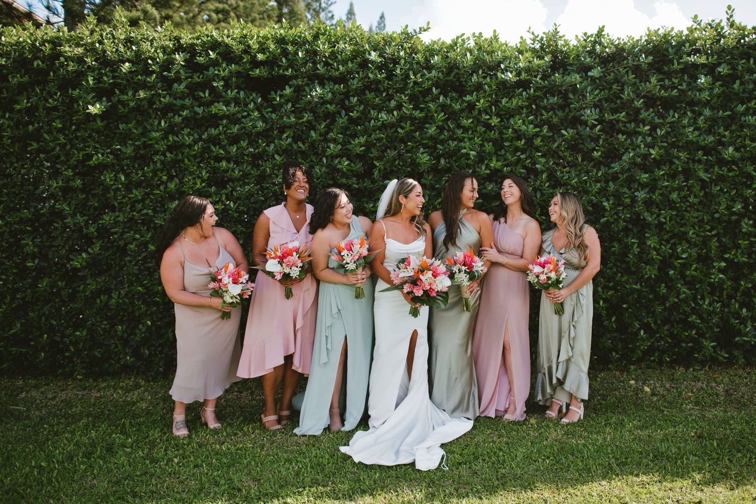 Bride in white gown stands with bridesmaids in pink and sage dresses holding colorful bouquets before a lush green hedge.
