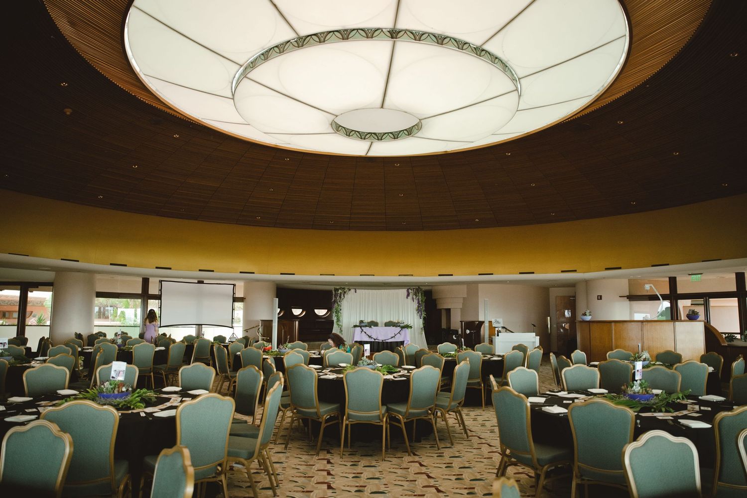 Elegant banquet hall with round ceiling light, green chairs, and set tables ready for a formal event.