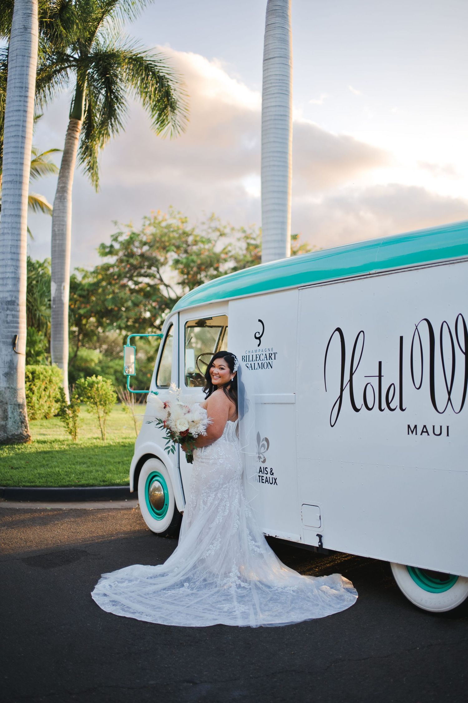 Bride in lace gown holding white bouquet poses beside a vintage Hotel Wailea Maui truck surrounded by palm trees.