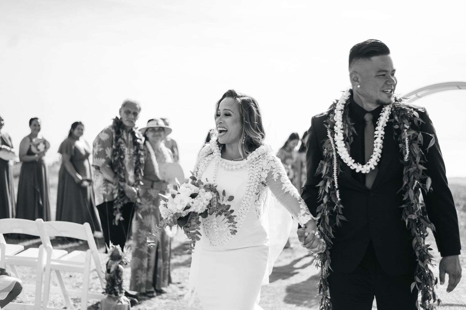 Black and white photo of a joyful bride and groom walking down the aisle at a beach wedding ceremony.