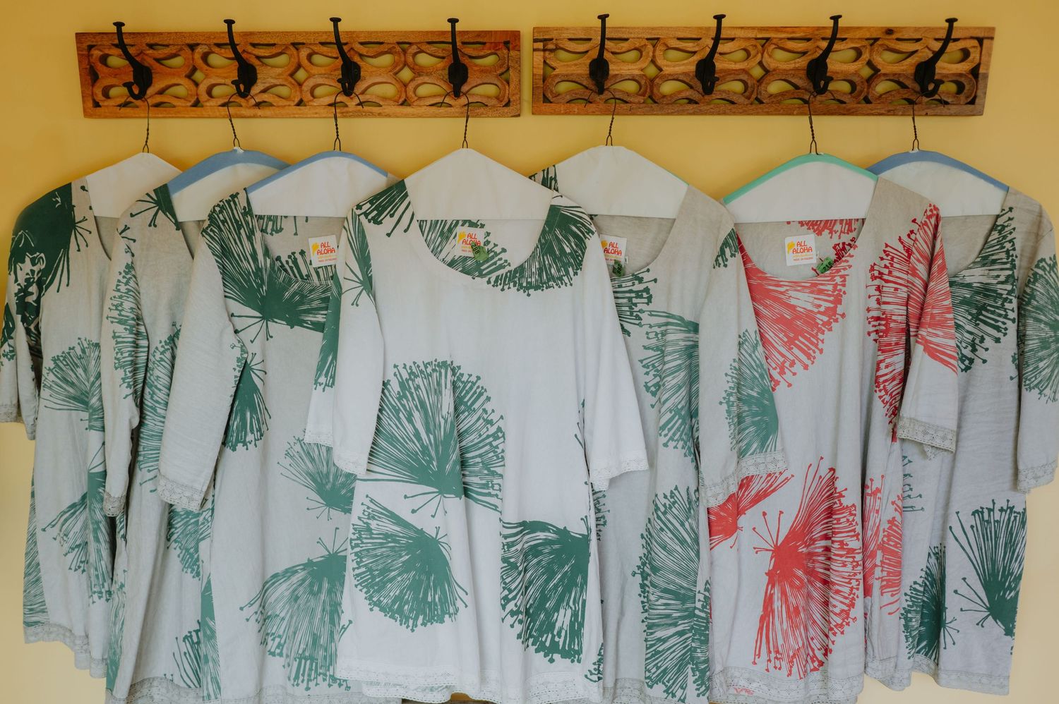 Colorful tropical leaf print shirts hanging on wooden wall hooks, featuring green and coral botanical patterns.