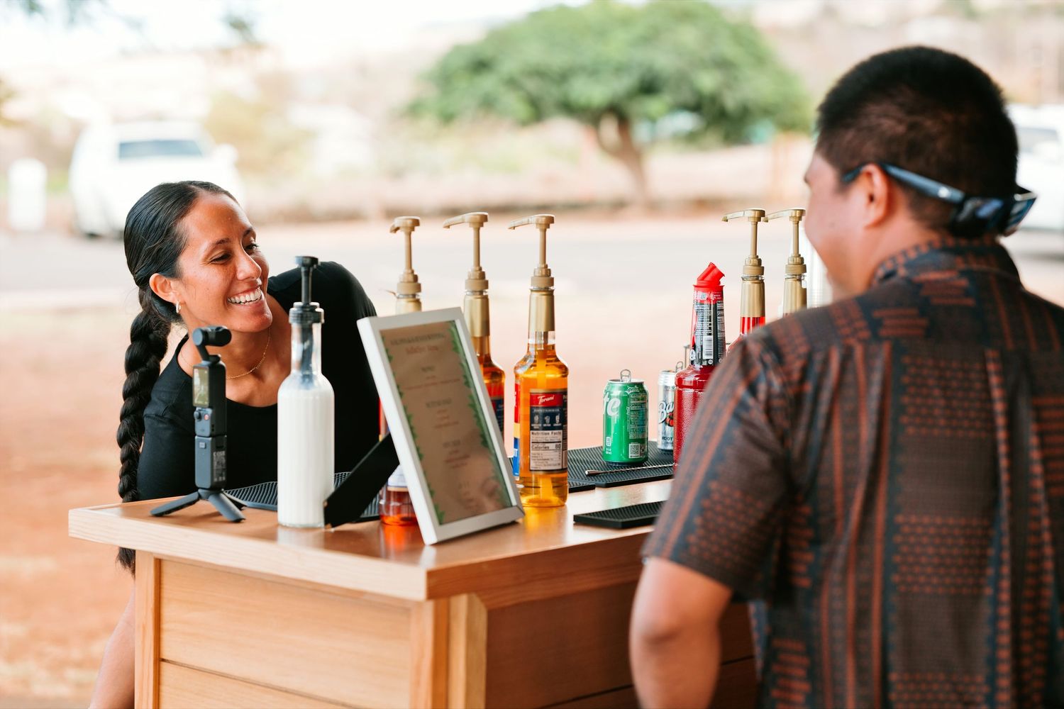 Woman smiling at a customer across an outdoor bar counter with bottles and a tablet display.