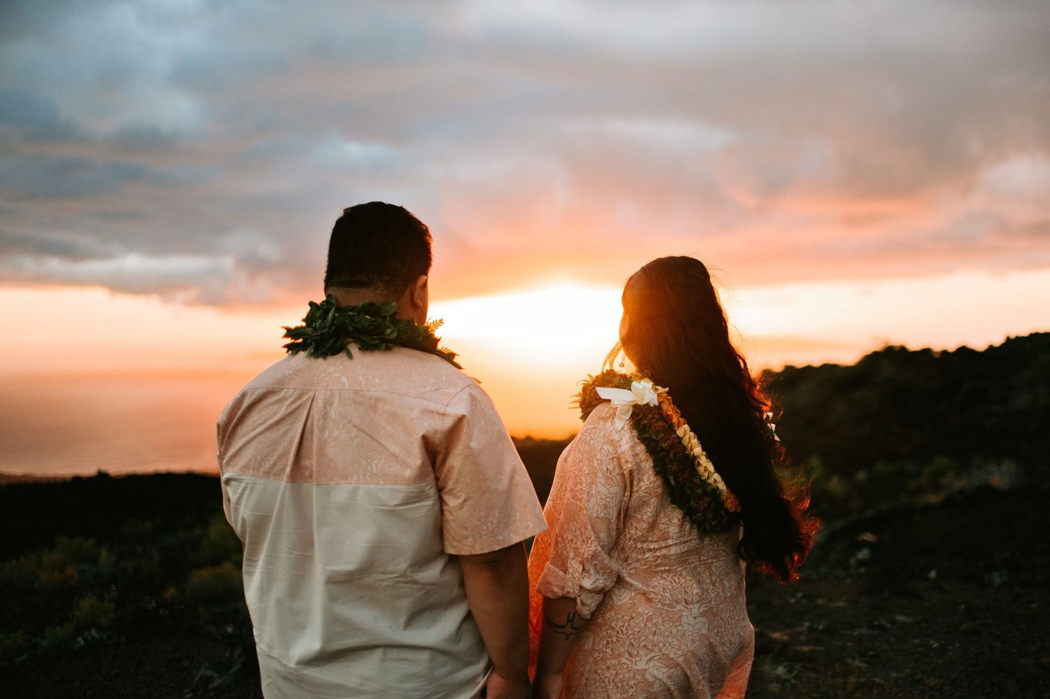 Couple holding hands watching a vibrant Hawaiian sunset, both wearing traditional lei garlands on rocky terrain.