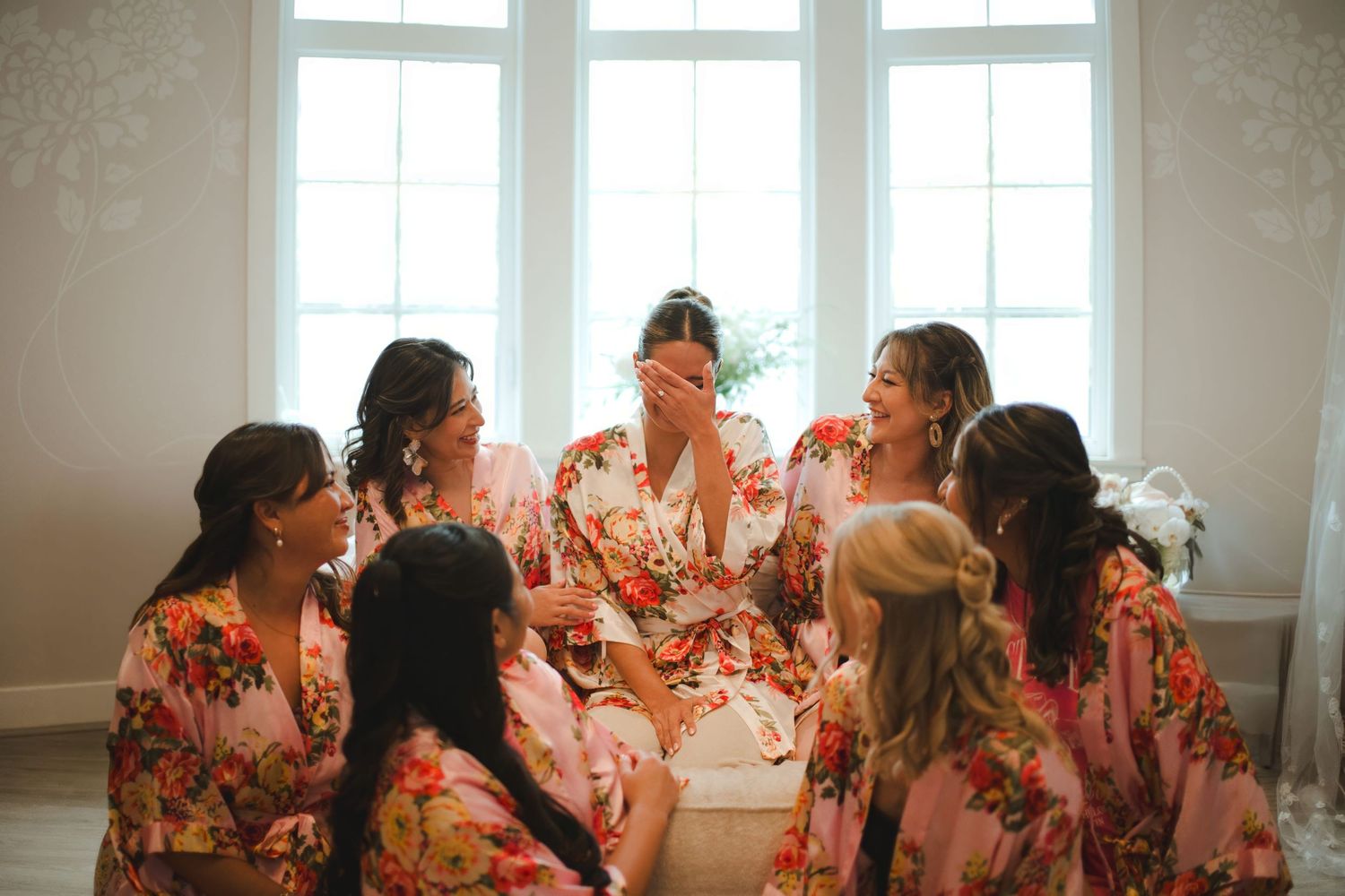 Bridesmaids laughing together in matching floral robes during wedding morning preparations near a bright window.