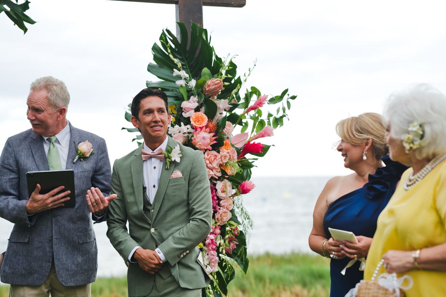 Groom in green suit smiles at outdoor beach wedding ceremony with tropical floral arch and ocean backdrop.