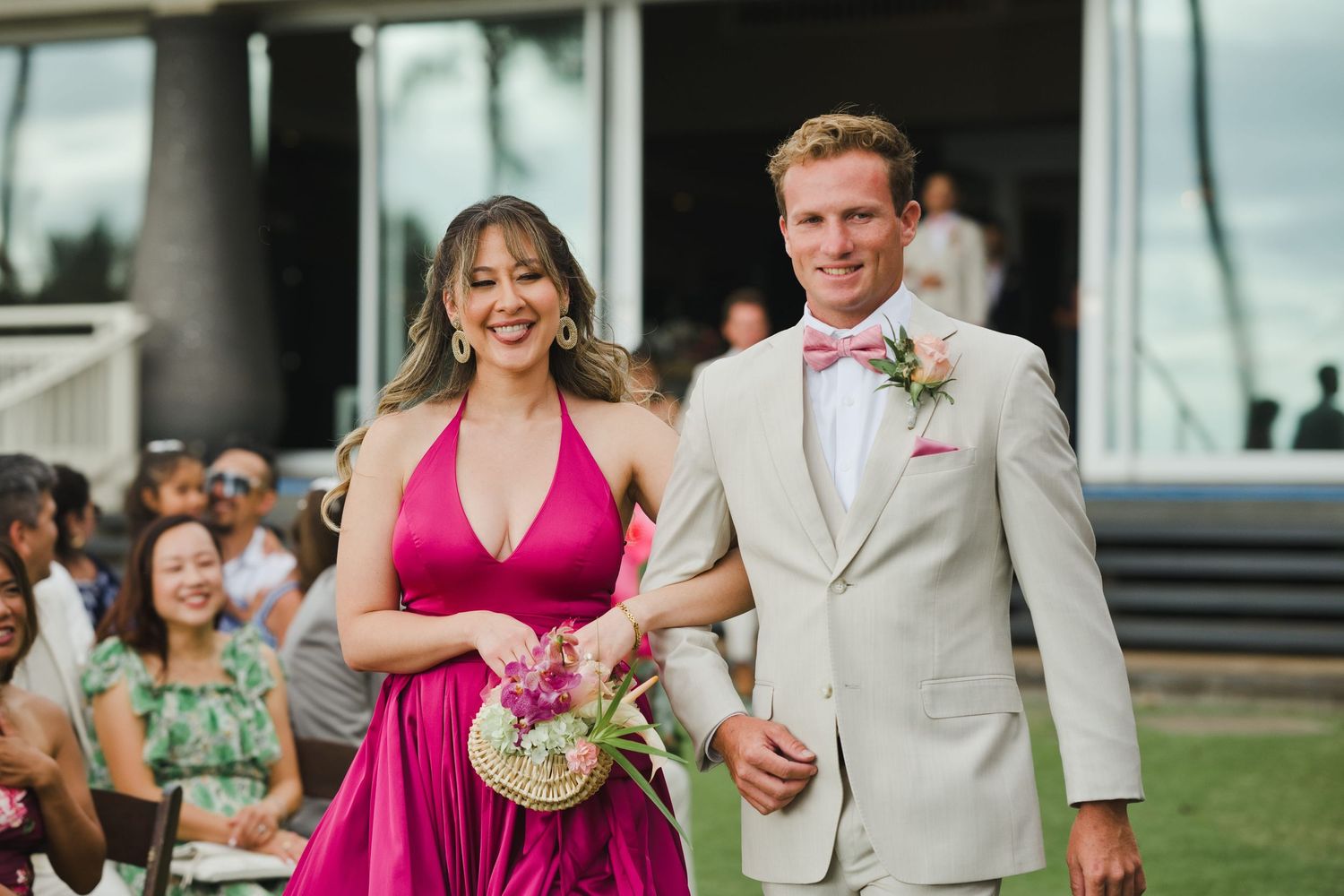 A smiling couple walks down the aisle at an outdoor wedding, woman in pink dress, man in tan suit with pink bow tie.