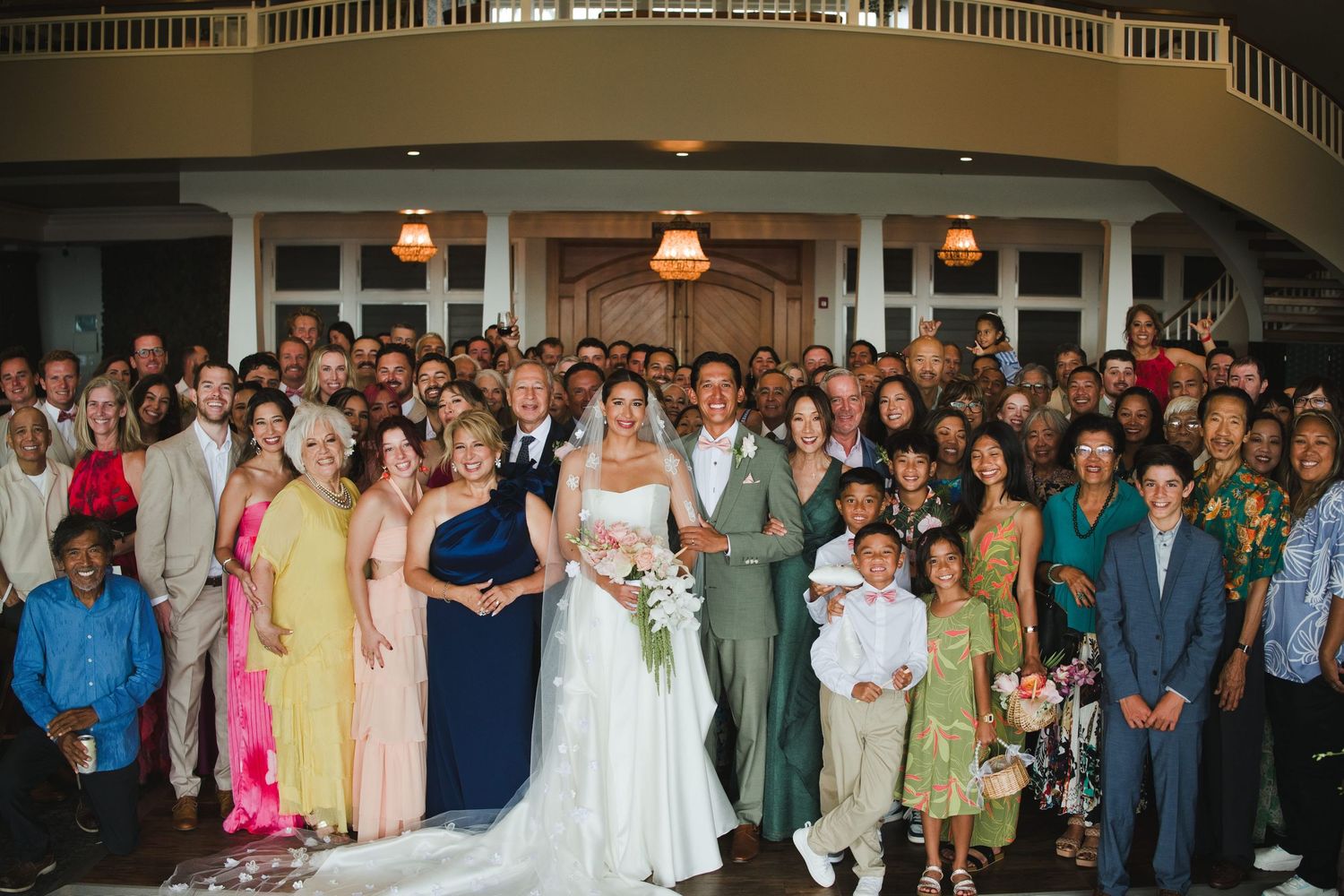 Large wedding group photo with bride and groom surrounded by guests in elegant venue.