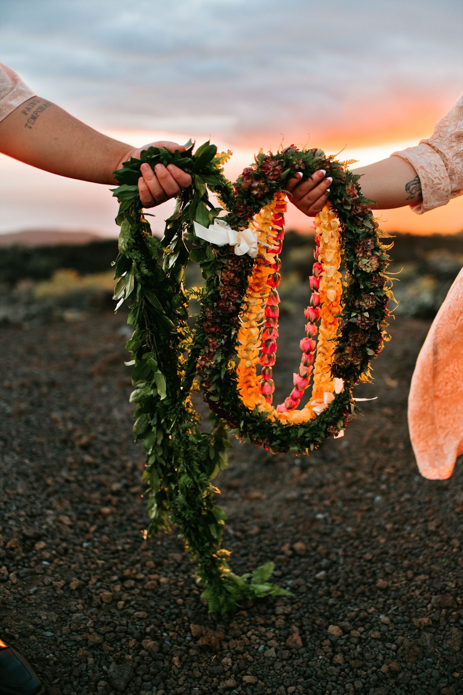 Two people hold colorful Hawaiian leis at sunset, featuring green maile and orange flower garlands against a volcanic landscape.