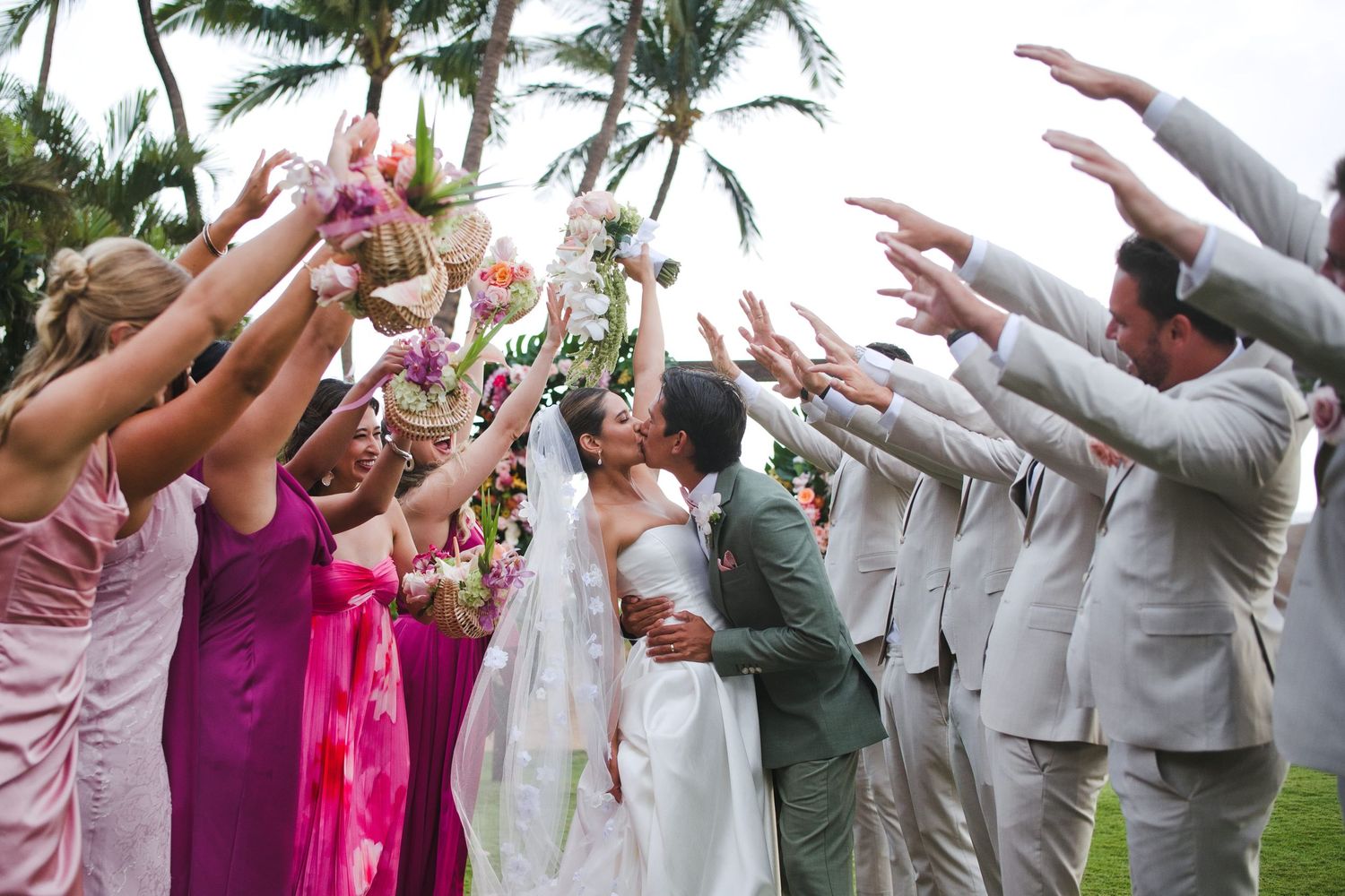Bride and groom kiss under an arch of raised arms formed by bridesmaids in pink and groomsmen in gray suits outdoors.
