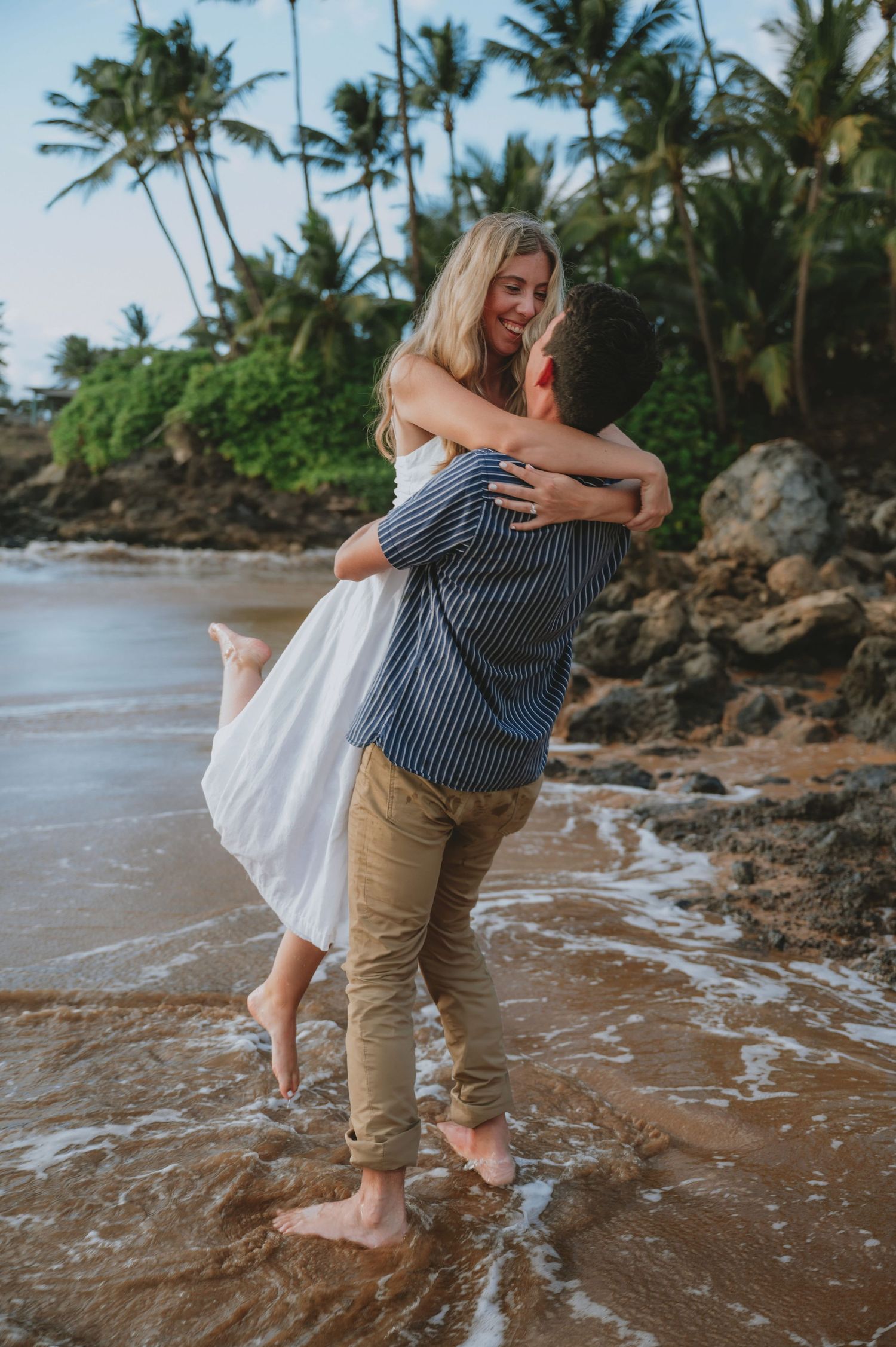 Couple embracing on a tropical beach, man lifting woman in white dress, palm trees and ocean waves in background.