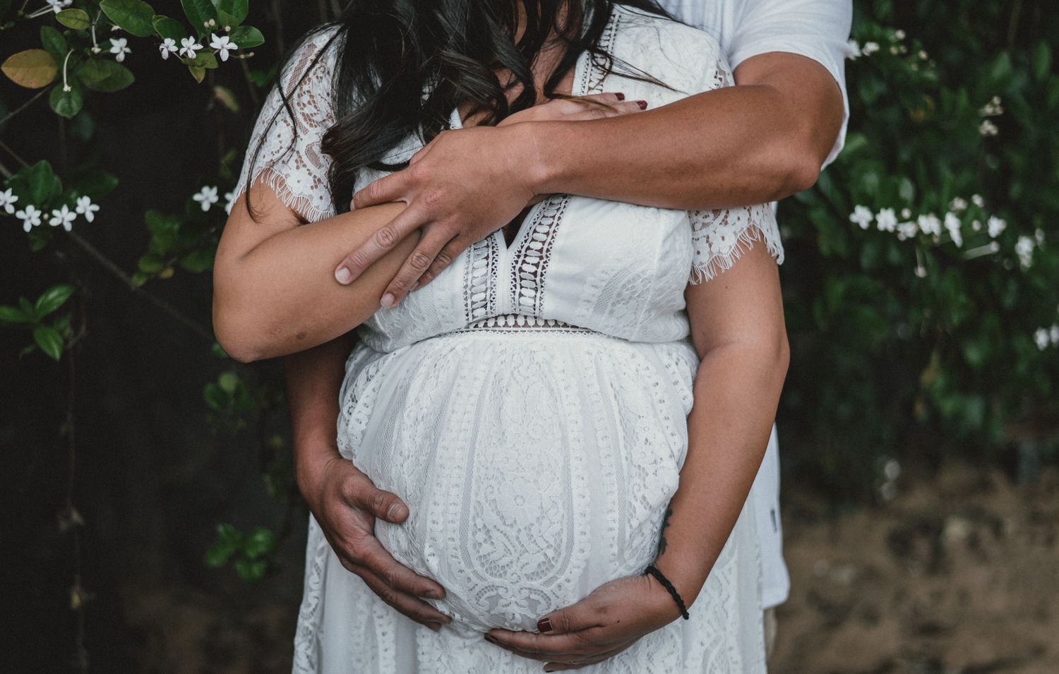 Close-up shot of hands cradling a pregnant belly in a white lace maternity dress against a natural backdrop.