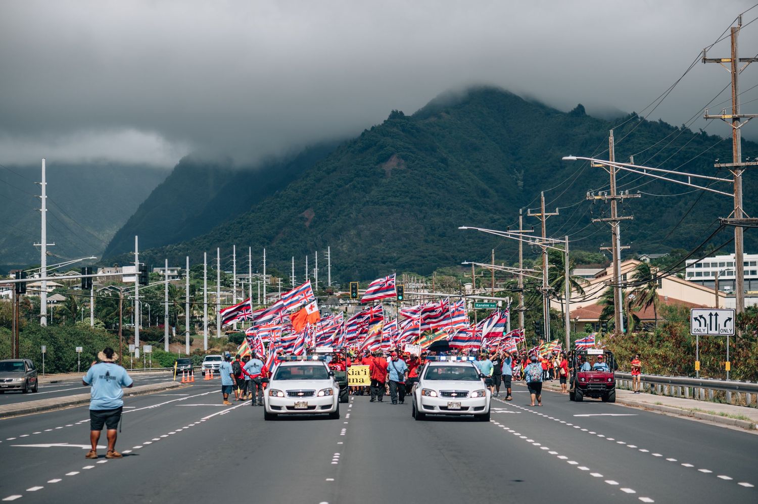 Large crowd with Hawaiian flags gathers along a road with mountains in the background.