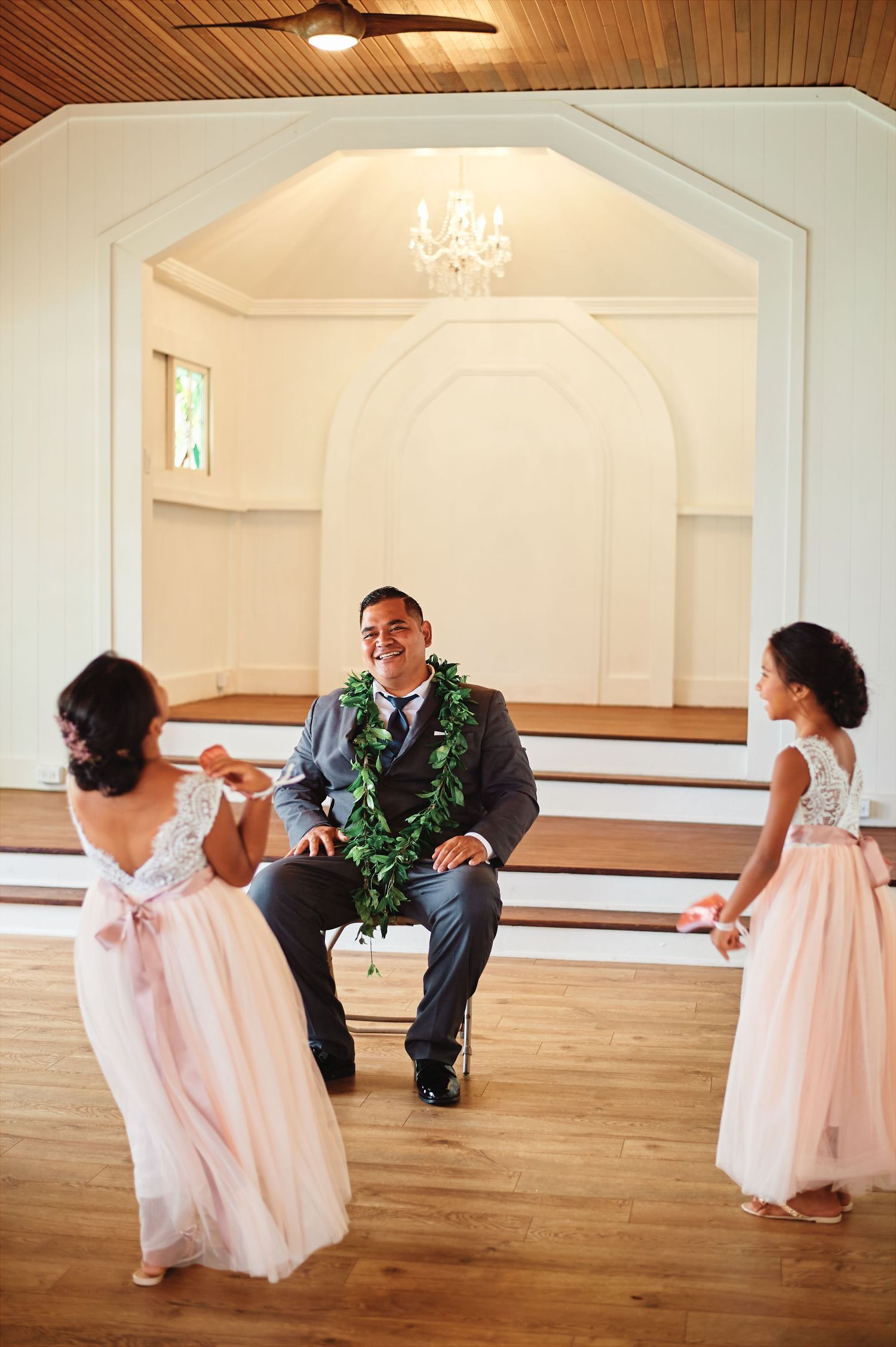 Wedding party members dance and play with children in formal attire in a bright reception hall.