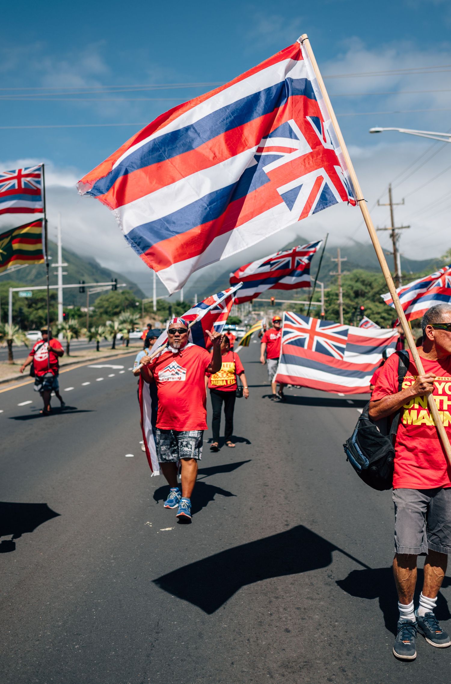 People waving Hawaiian state flags march along an asphalt road with mountains and blue sky in background.