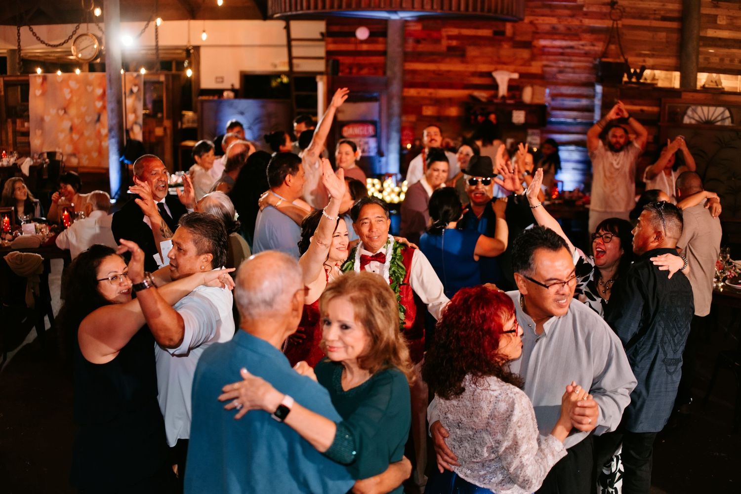 Guests dancing and celebrating at a lively indoor wedding reception in a rustic venue with warm lighting.