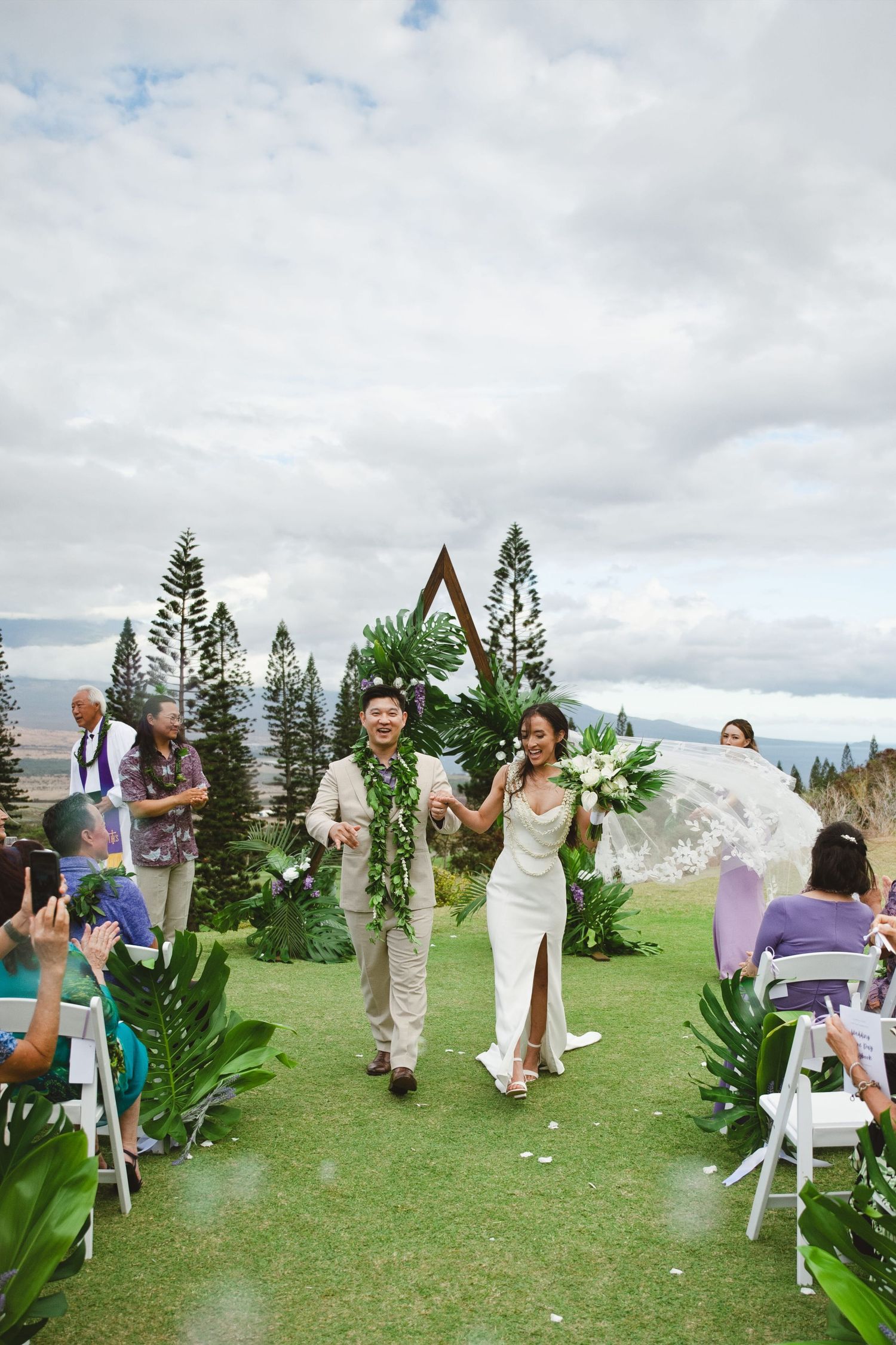 Newlywed couple walks down outdoor aisle at tropical Hawaii wedding ceremony with lush greenery and geometric arch backdrop.