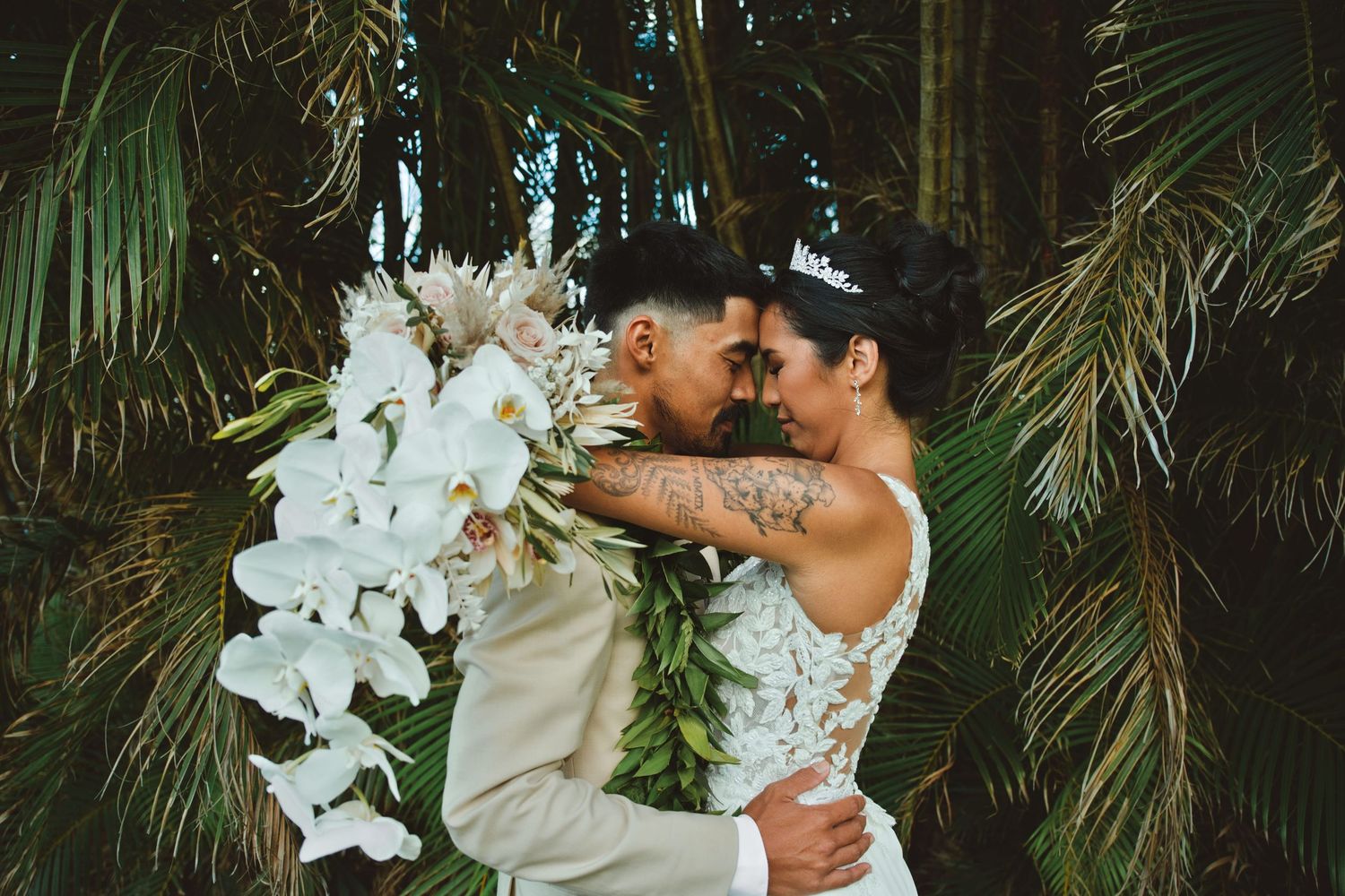 Bride and groom share romantic moment surrounded by tropical palms, bride holding white orchid bouquet.