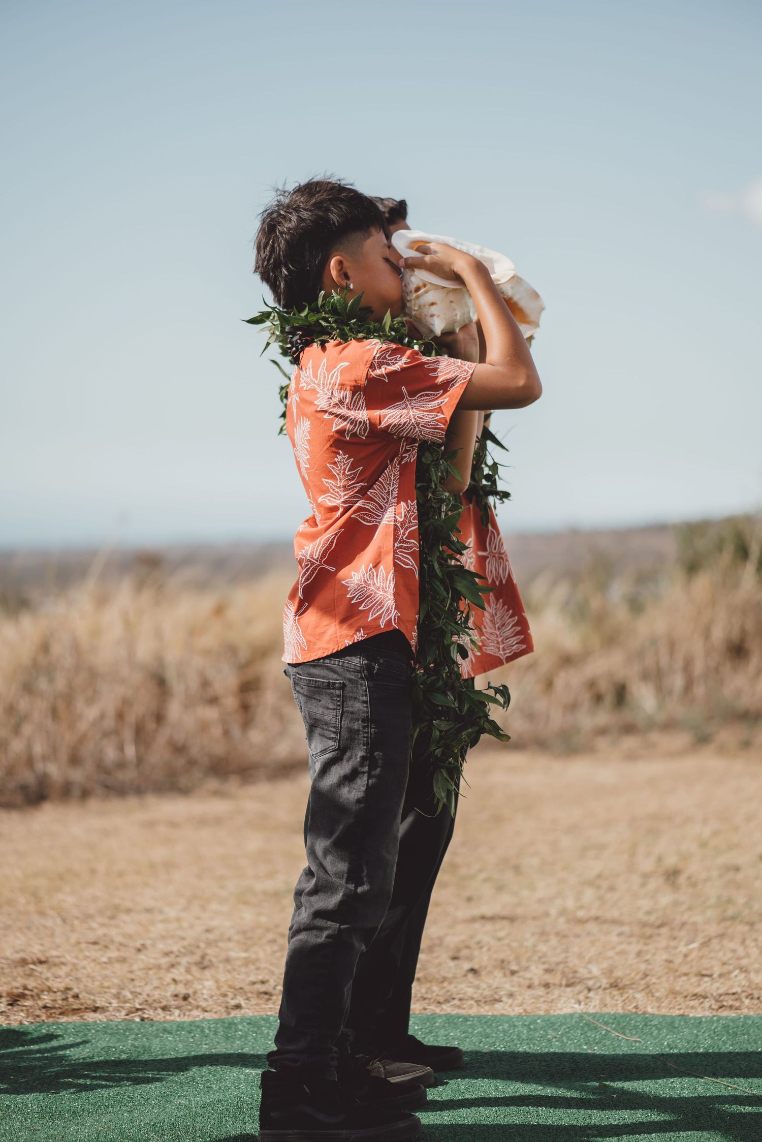 Boy in Hawaiian shirt and lei blowing a conch shell outdoors in Hawaii.