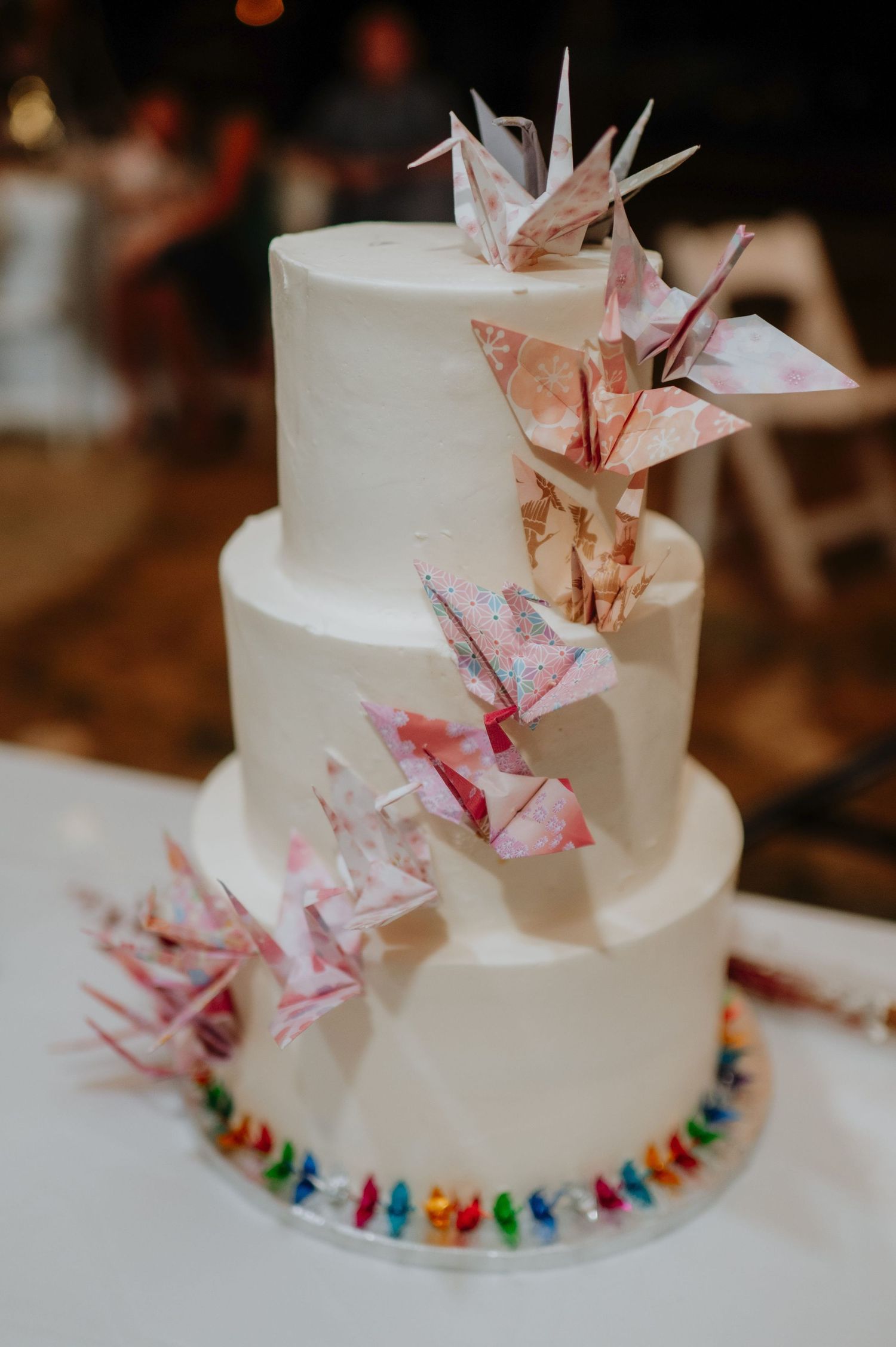 Three-tier white wedding cake decorated with colorful origami paper cranes cascading down the layers.