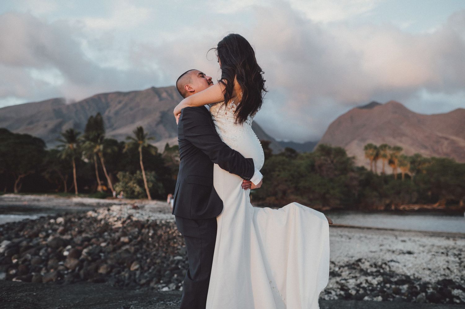 Couple in wedding attire embracing on rocky beach with tropical mountains and palm trees in background.