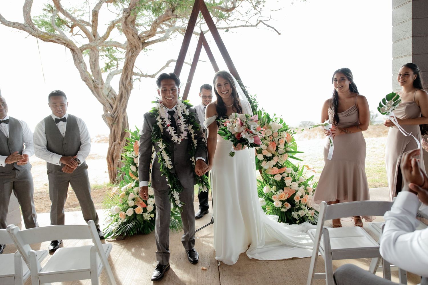 Bride and groom walk down the aisle at an outdoor wedding ceremony with a floral triangular arch backdrop.