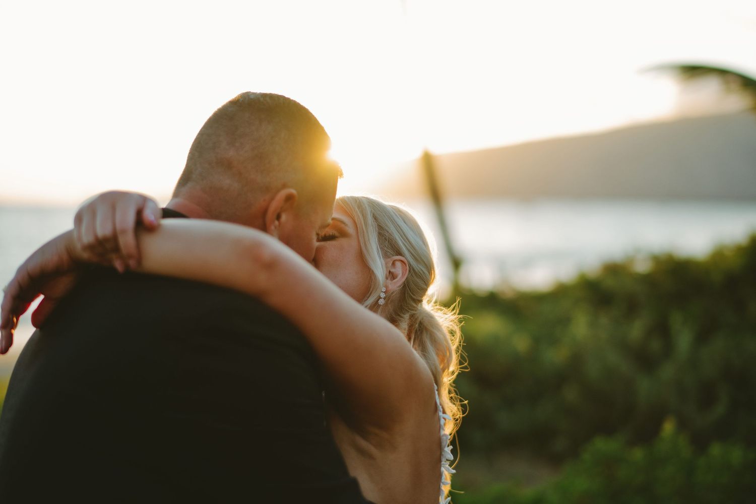 Couple sharing a romantic kiss at sunset with ocean and lush greenery in the background.