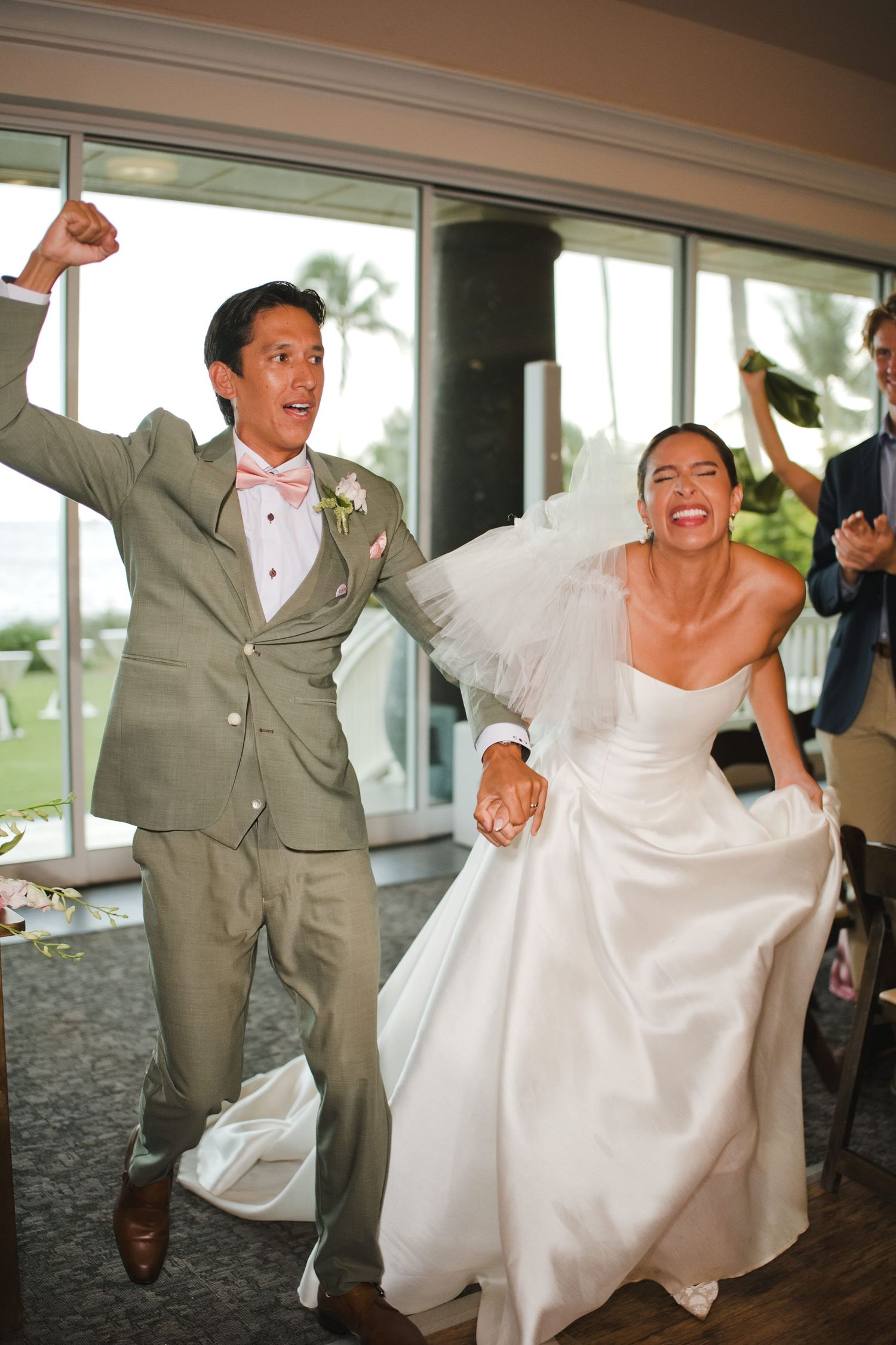 Joyful bride and groom make grand entrance at wedding reception, laughing and cheering with hands raised.