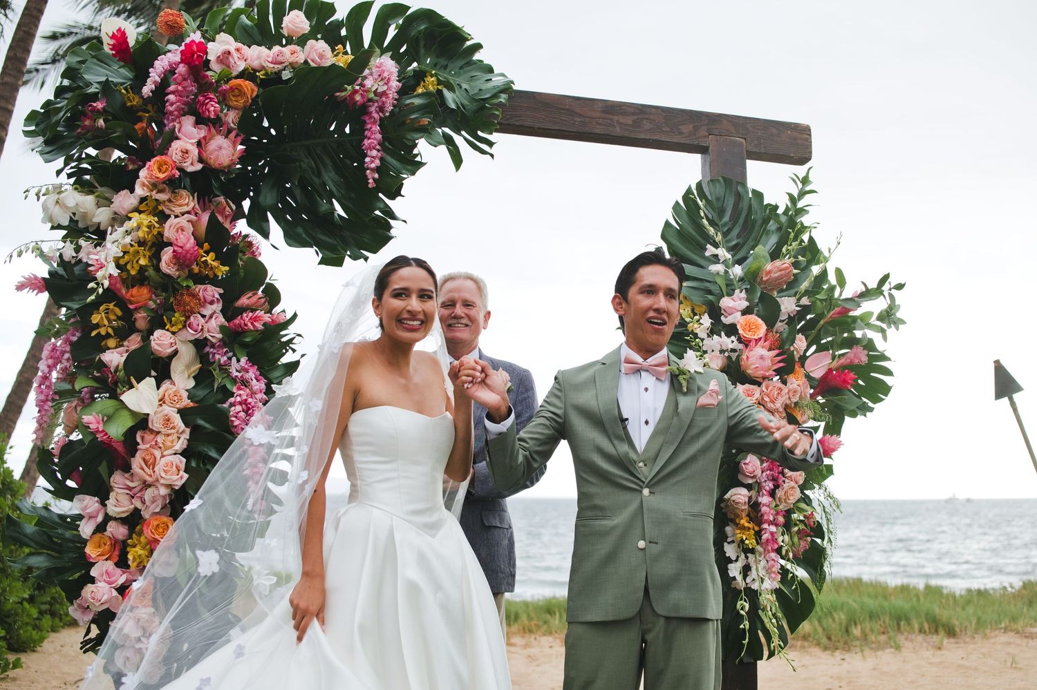 Newlywed couple celebrating at outdoor beach wedding ceremony under floral arch with colorful tropical flowers.