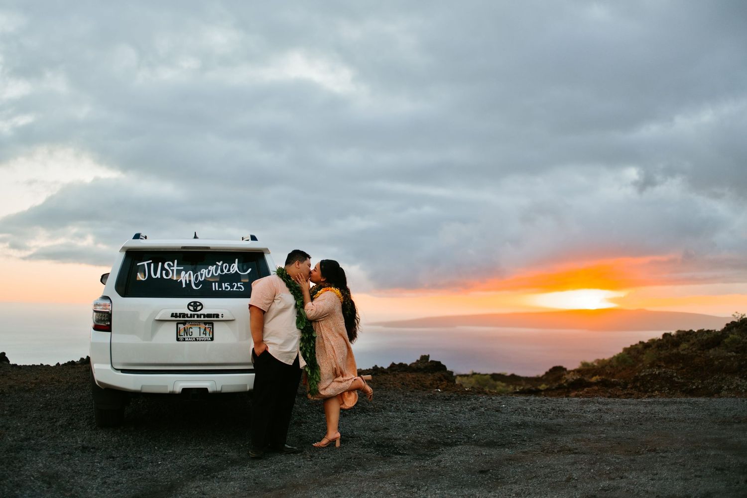 Newlywed couple kissing beside a white SUV with Just Married sign at sunset on a rocky Hawaiian landscape.