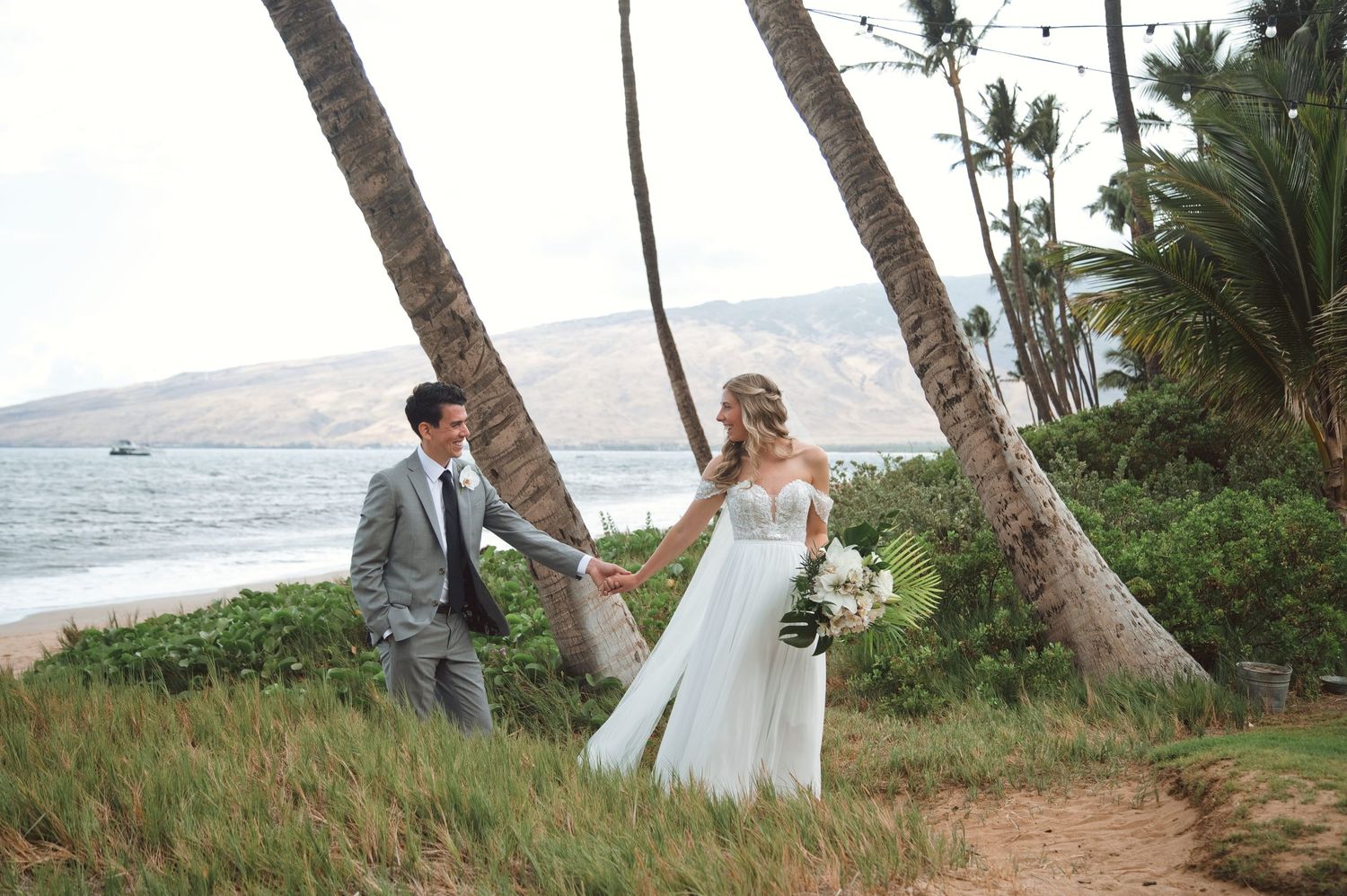 Bride and groom walk hand in hand on a tropical beach path lined with palm trees in Maui, Hawaii.