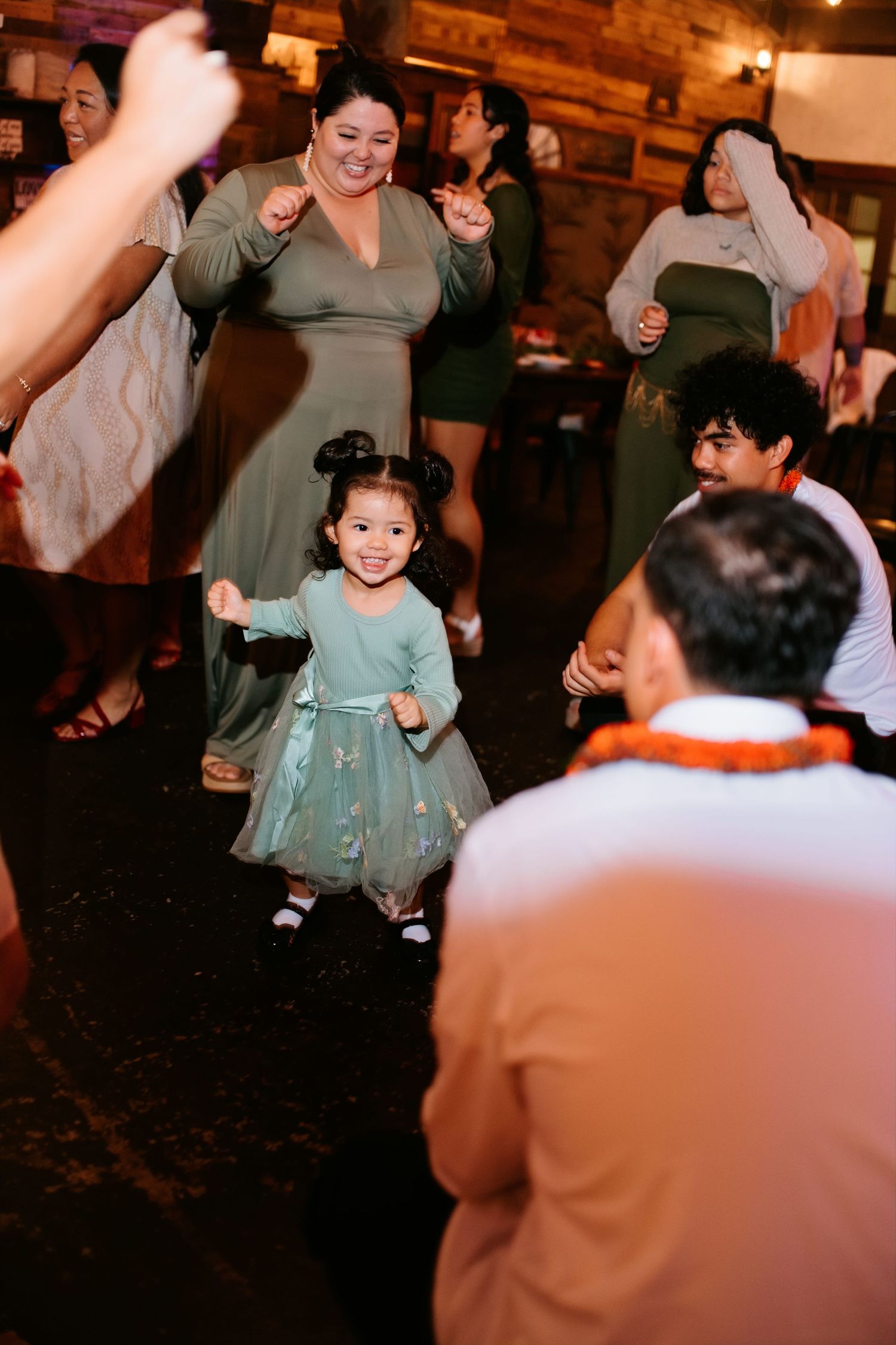 A joyful little girl in a teal dress dances at a wedding reception surrounded by guests in green dresses.