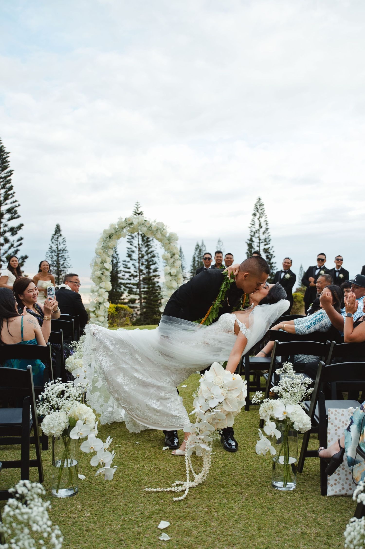 Groom dips bride for a kiss at outdoor wedding ceremony with white floral arch and aisle decorations.