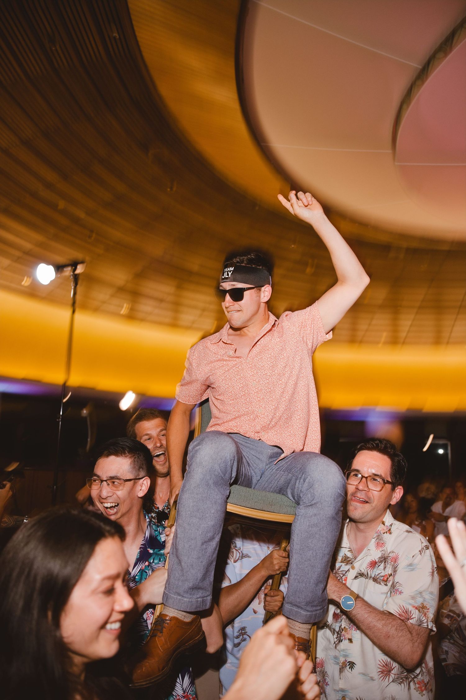A young man in a pink polo and sunglasses is lifted by a crowd at a lively party under a domed ceiling.