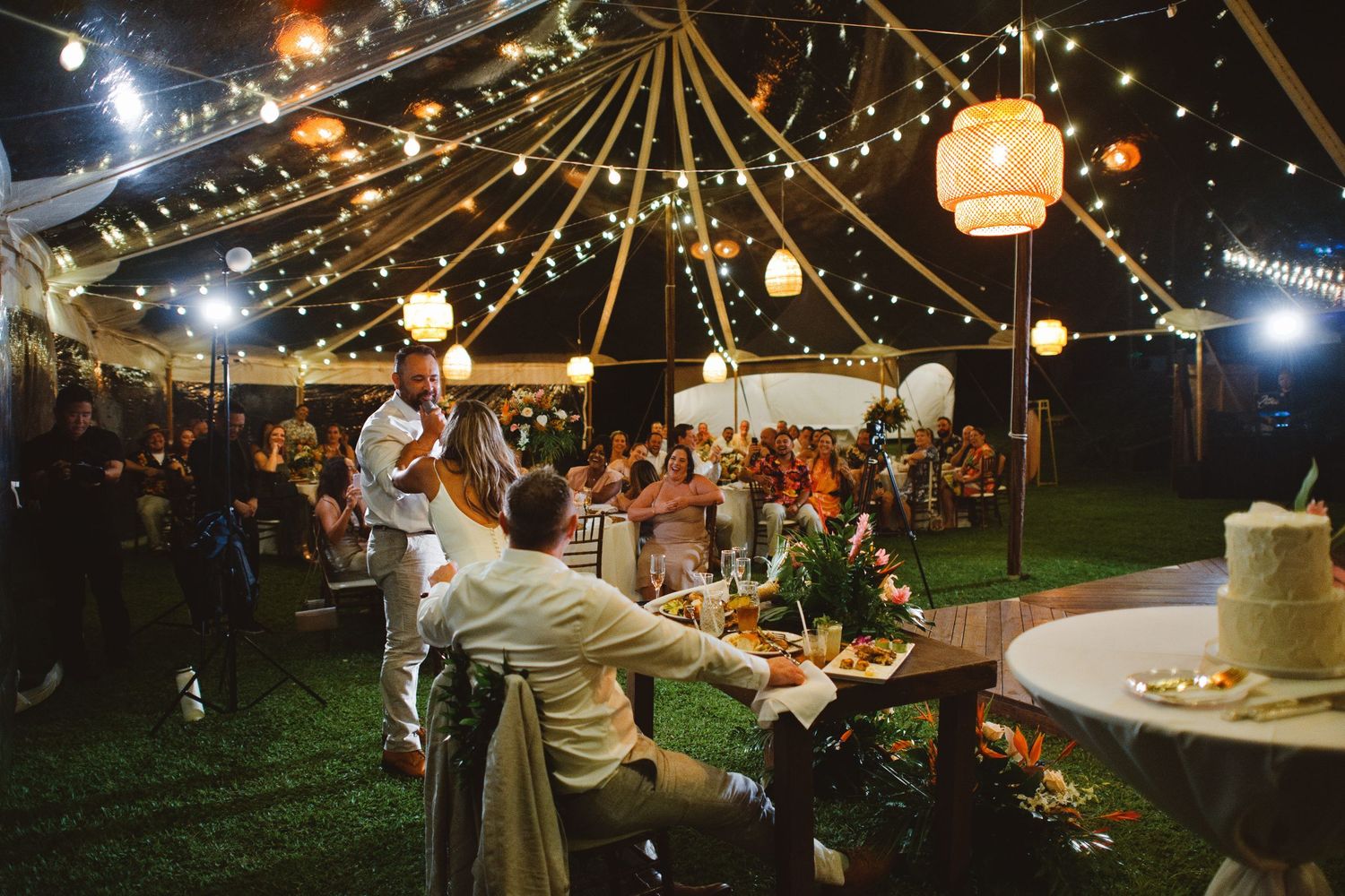 Outdoor evening wedding reception under string lights and lanterns with guests dining on green grass.