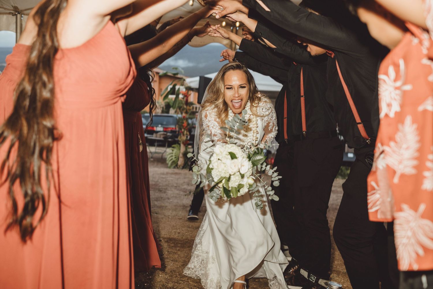 Bride laughing joyfully while walking through a tunnel of raised hands formed by wedding guests outdoors.
