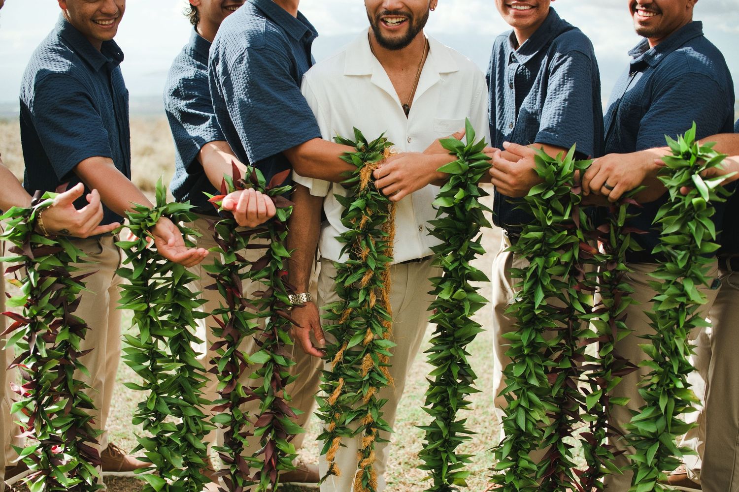 Groom in white shirt stands with groomsmen holding green leaf leis on a Hawaiian beach wedding.