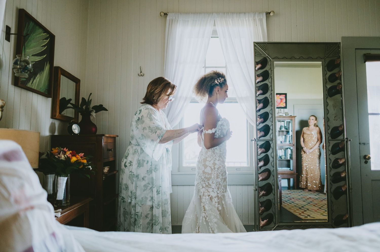 Bride being helped into her lace wedding gown by a woman in a floral dress, reflected in a full-length mirror.