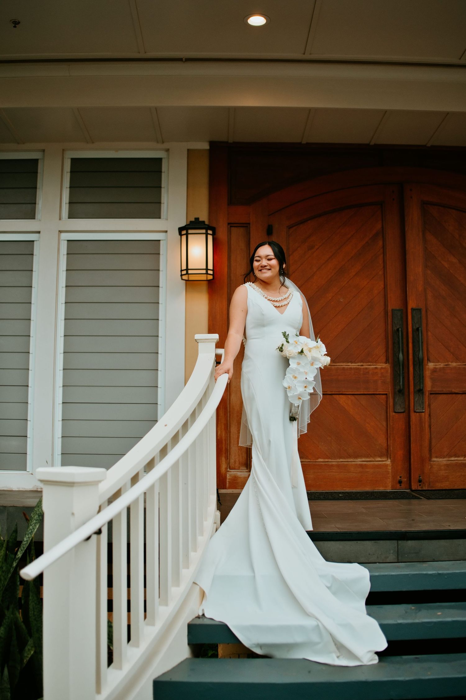 Bride in white gown holding bouquet stands on steps before wooden doors.