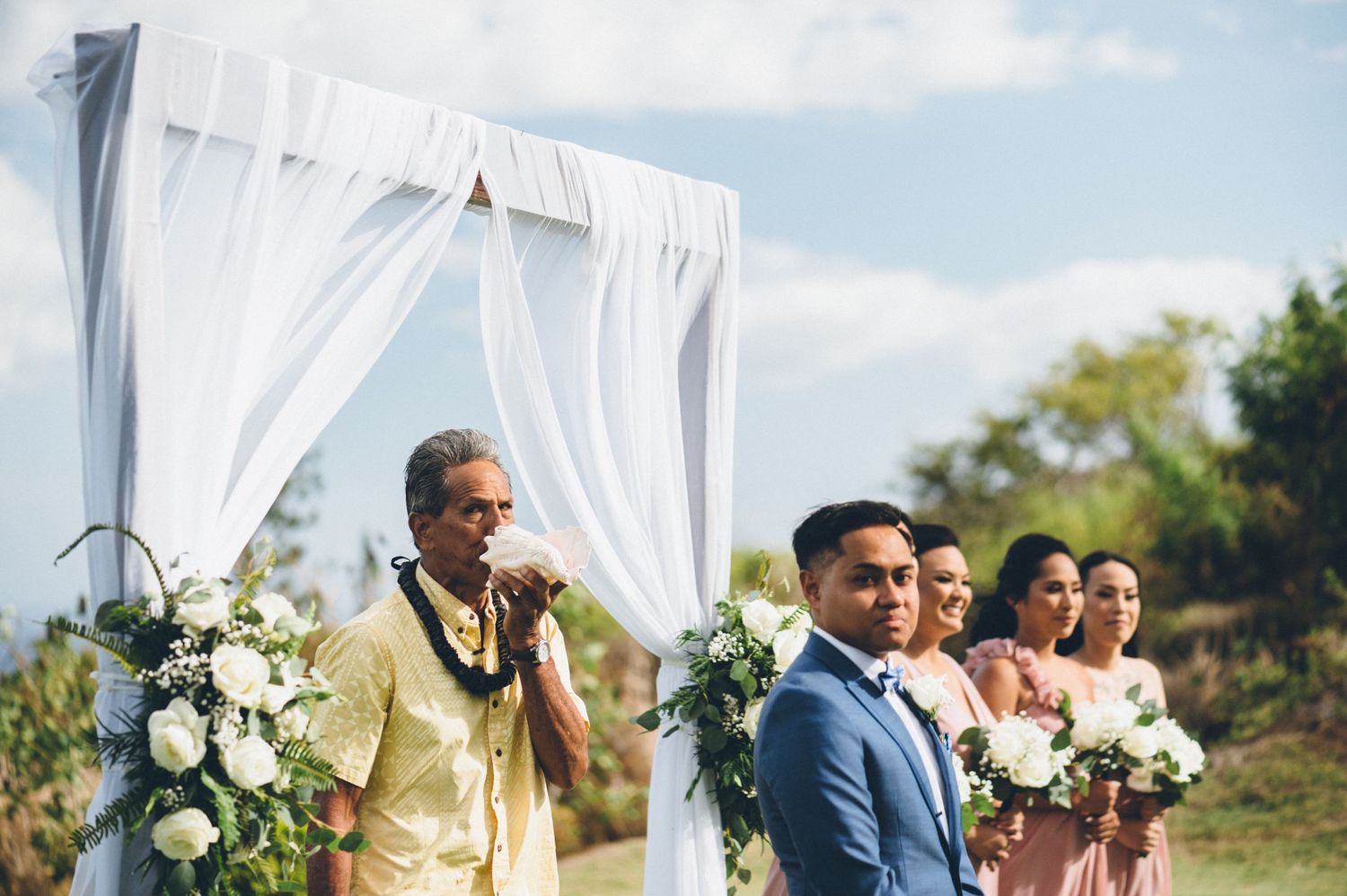 Outdoor wedding ceremony setup with white draped arch and wedding party in formal attire.