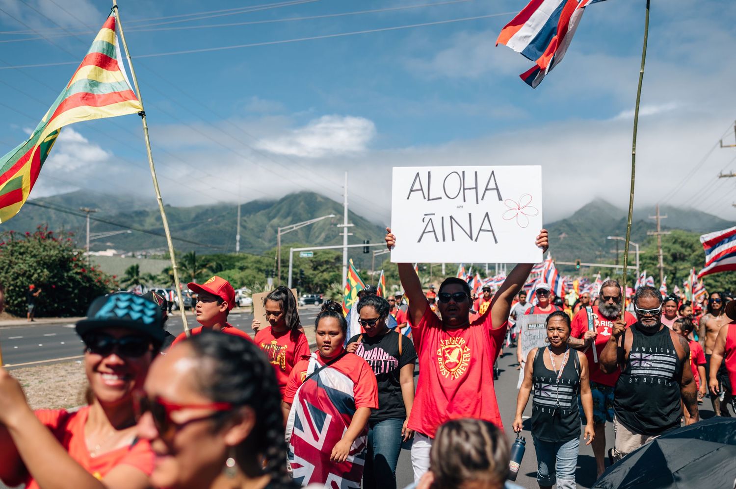 Protesters carry Aloha and Aina signs while marching with Hawaiian flags during demonstration.