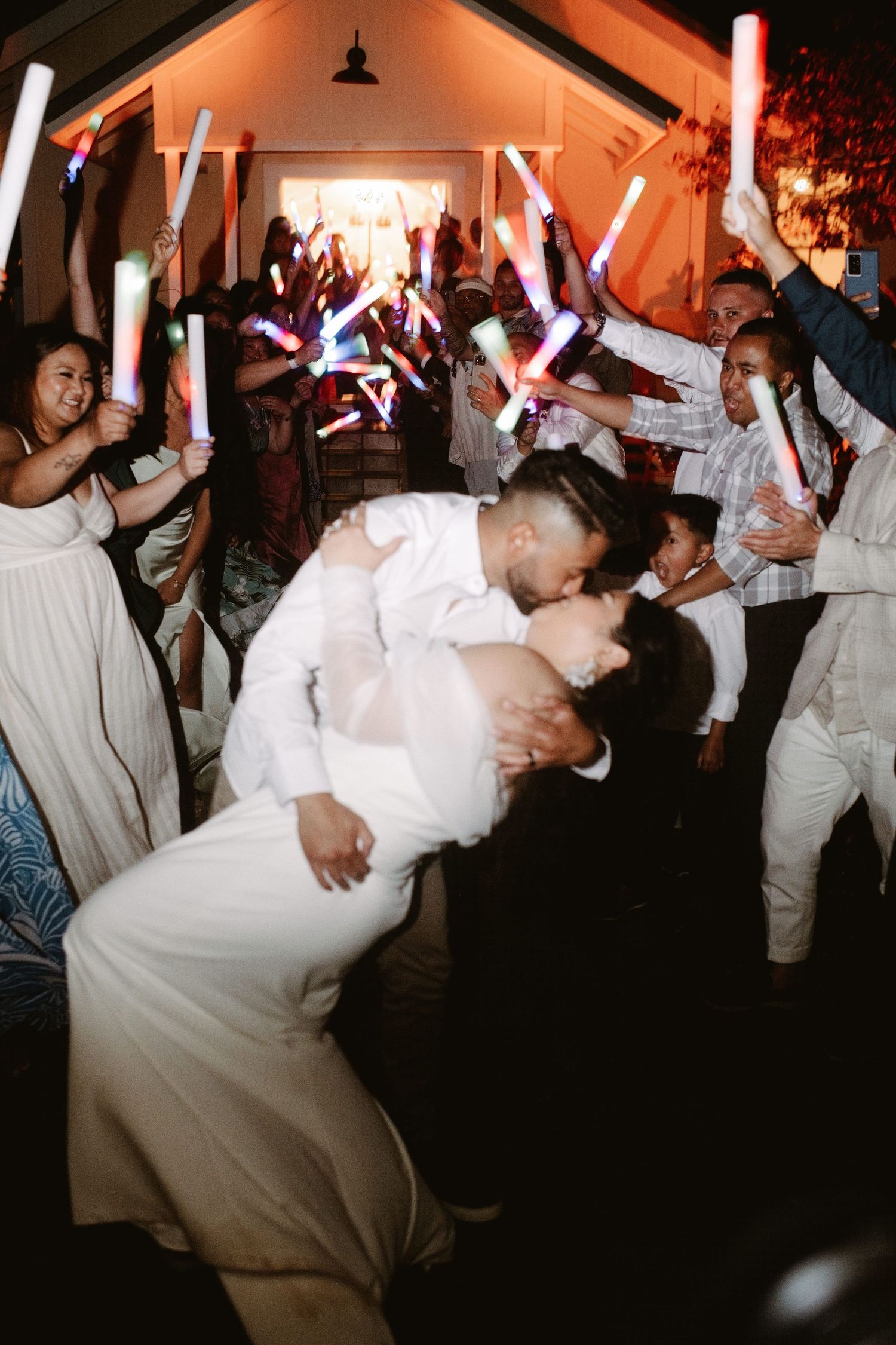 Newlyweds share a dip kiss surrounded by cheering guests waving colorful glow sticks at a festive wedding reception.