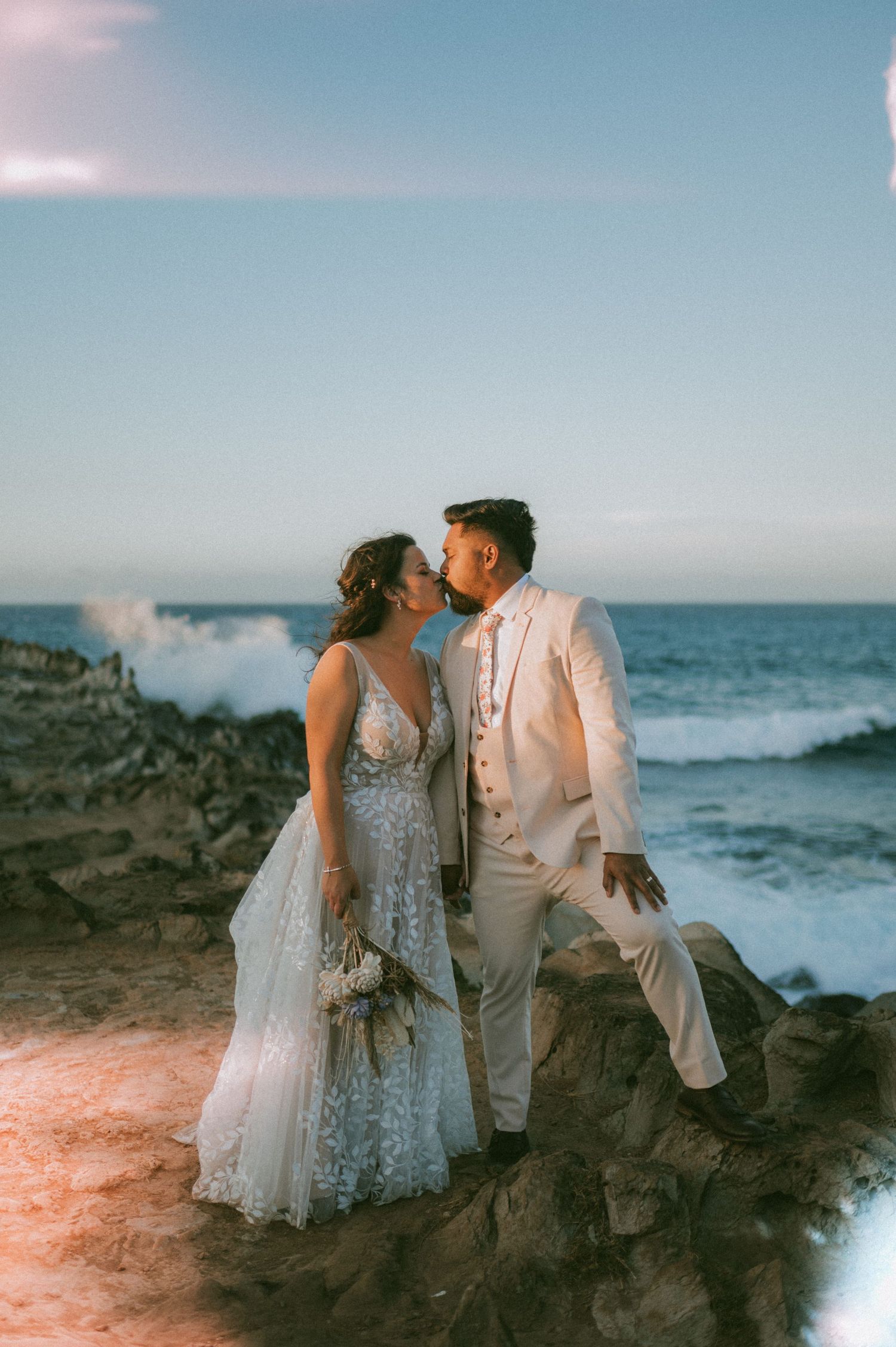 Wedding couple shares a romantic kiss on rocky oceanside cliffs at sunset with crashing waves behind them.